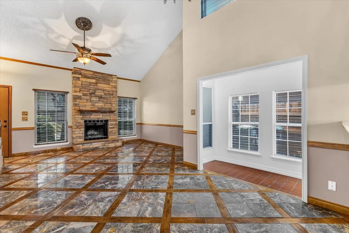 Vaulted ceiling, stone-clad fireplace with hearth, wood-finish and tile flooring, ceiling fan with light fixture, and multiple windows. Soaring ceilings with transom windows, opens into the sun room.