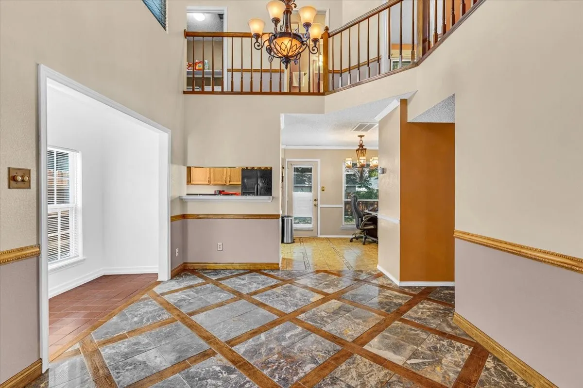 This living room view features soaring ceilings with transom windows, a stone-clad fireplace, and an elegant chandelier. open to the Dining Room and Kitchen.