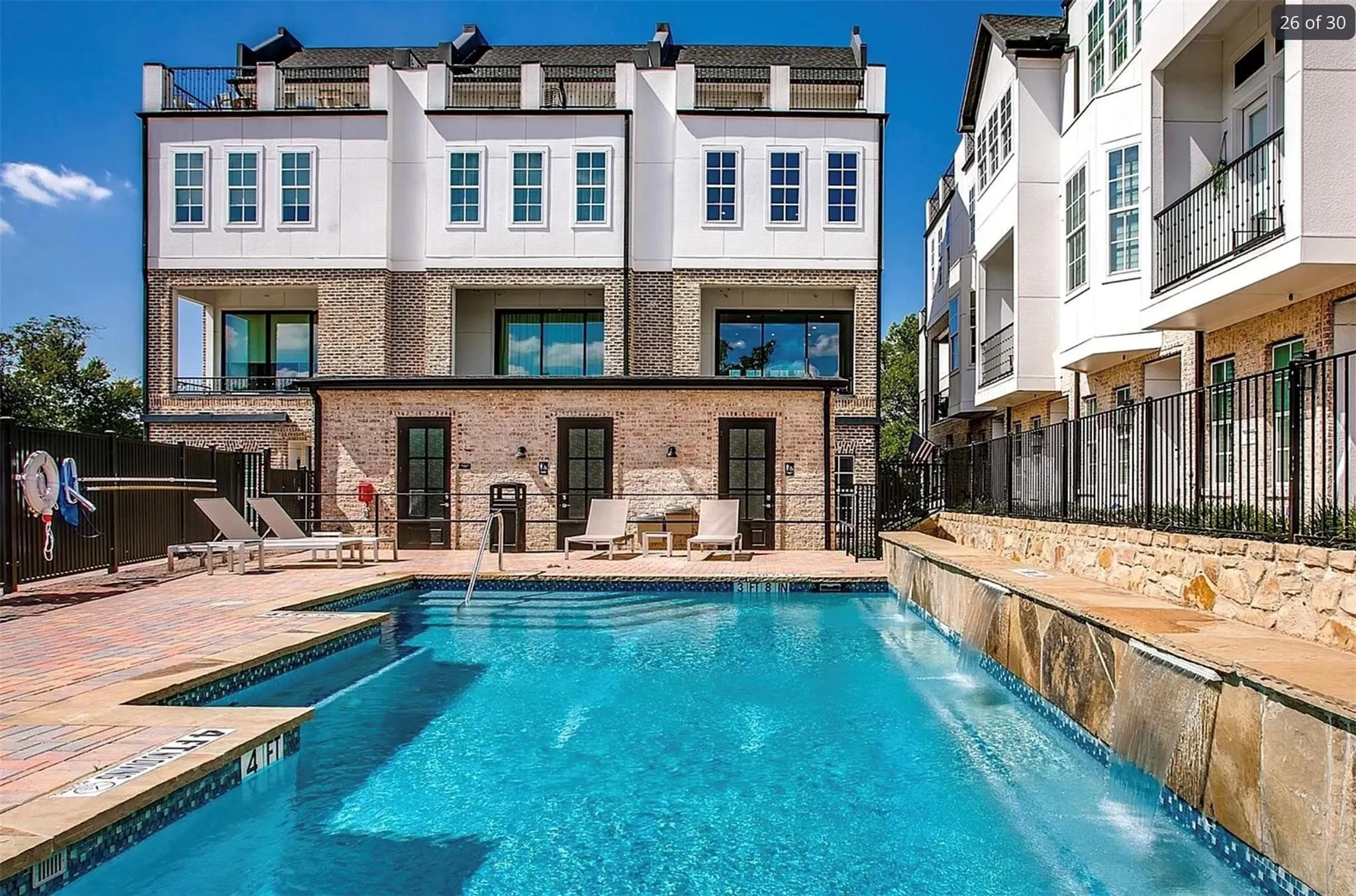 Outdoor swimming pool with integrated stone waterfall feature, brick paver decking, and metal privacy fencing
