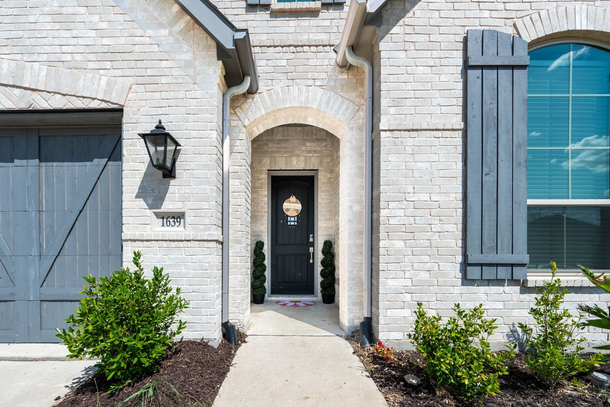 White brick exterior, arched entryway, dark wood front door with decorative glass, arched window with wood shutters, and dark wood-style garage door