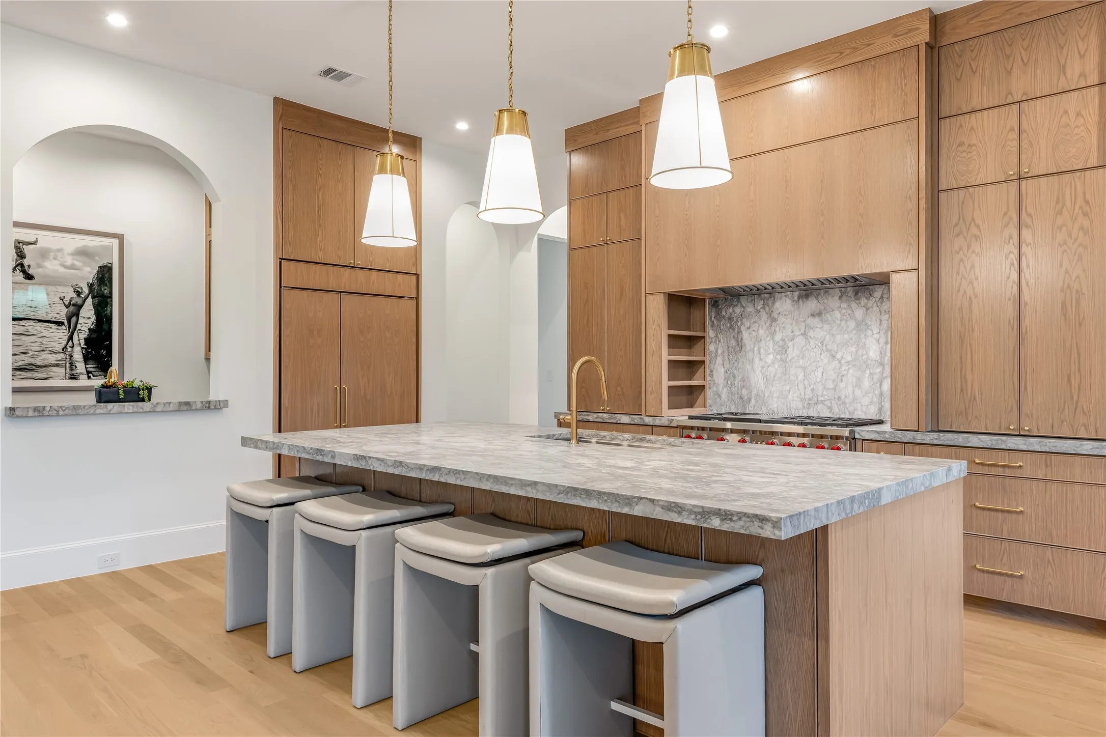 Center island with a stone countertop, three pendant lights, wood-finish flooring, full-height wood cabinetry, and an arched wall opening