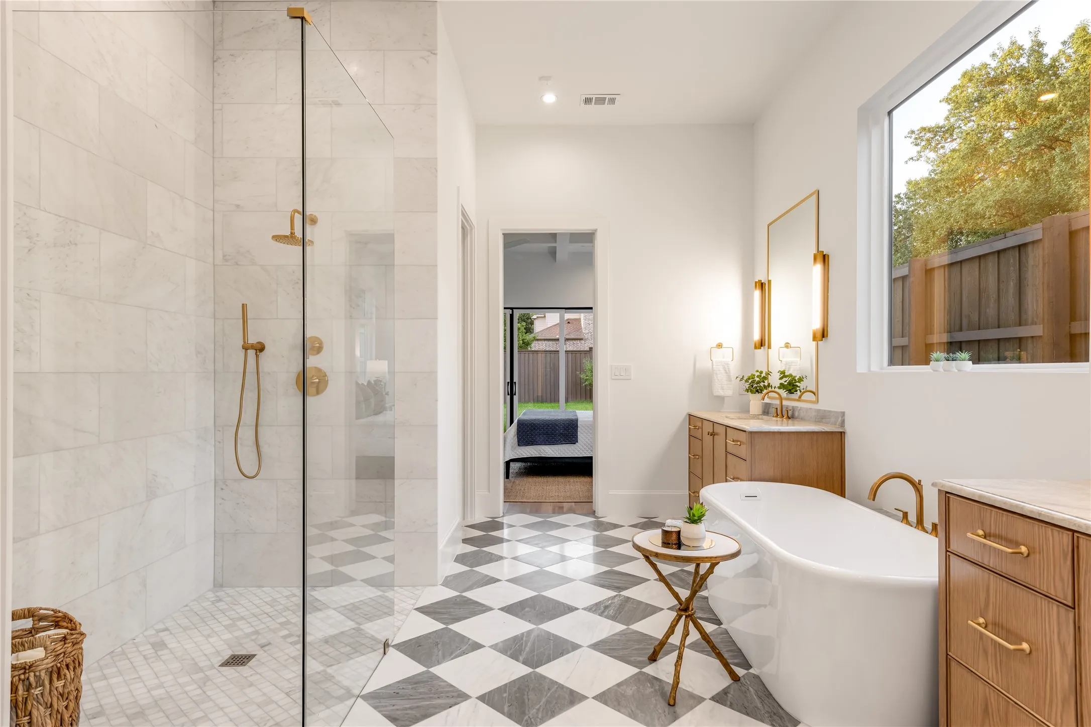 Glass-enclosed shower with gold-tone fixtures, freestanding soaking tub, wood-finish vanity with stone countertop, checkerboard tile flooring, and a large window