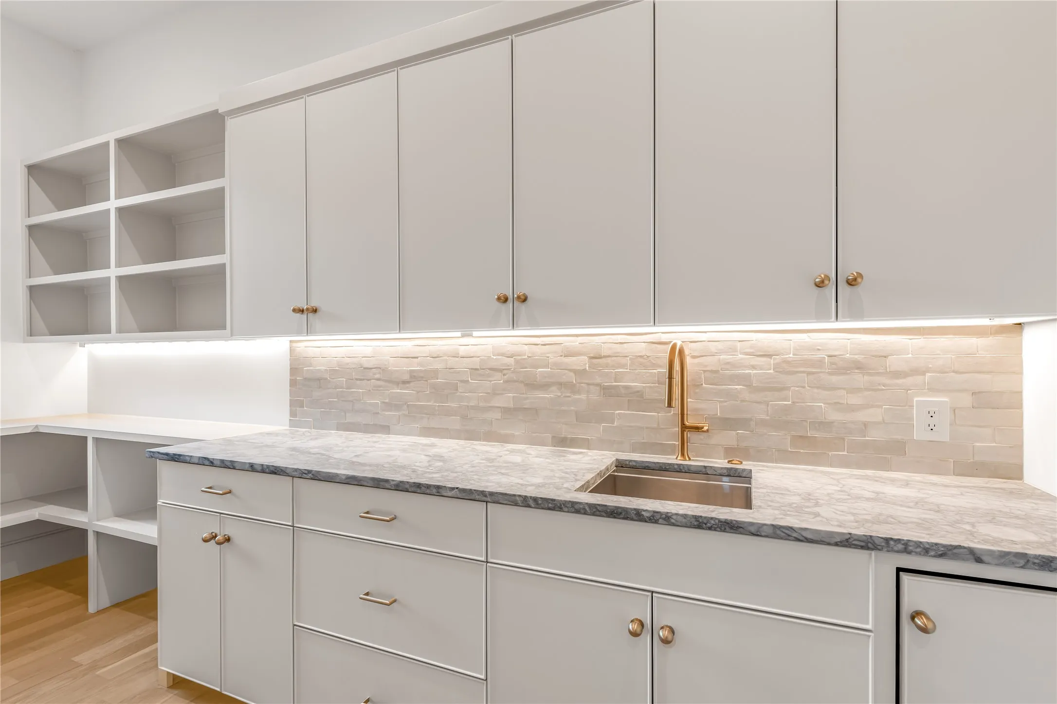 Built-in cabinetry with brass hardware, undermount sink with brass faucet, stone-finish backsplash, and under-cabinet lighting