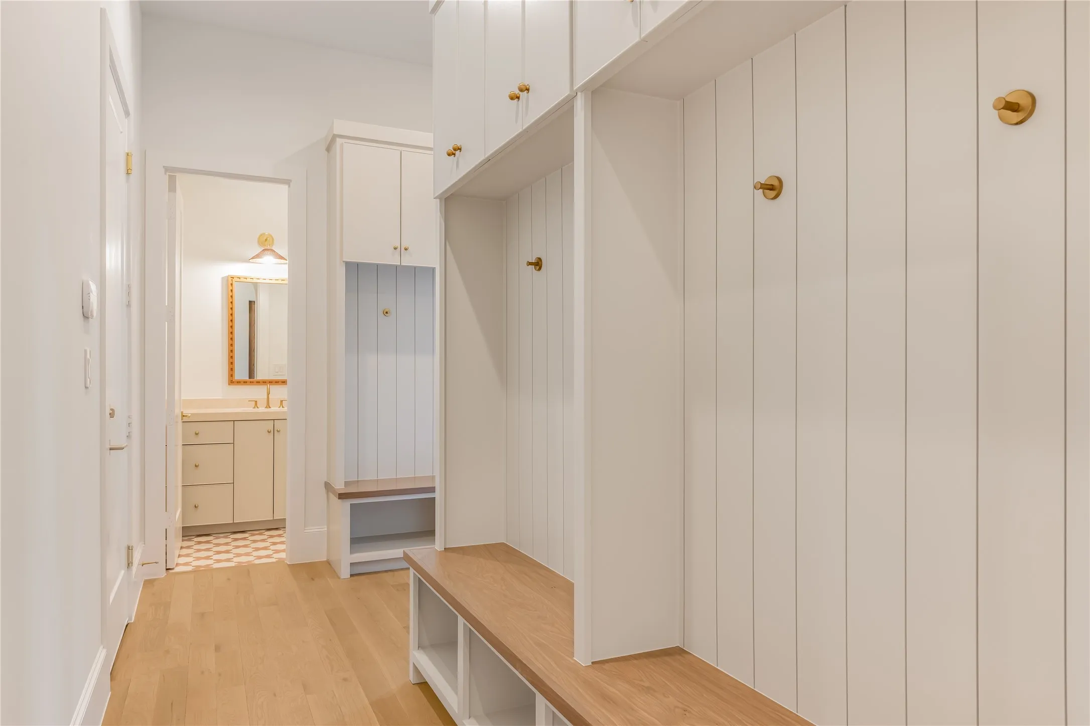 Built-in storage bench with wood-finish top and open shelving, vertical paneled wall accents, brass-finish hooks, overhead cabinetry, and light wood-finish flooring