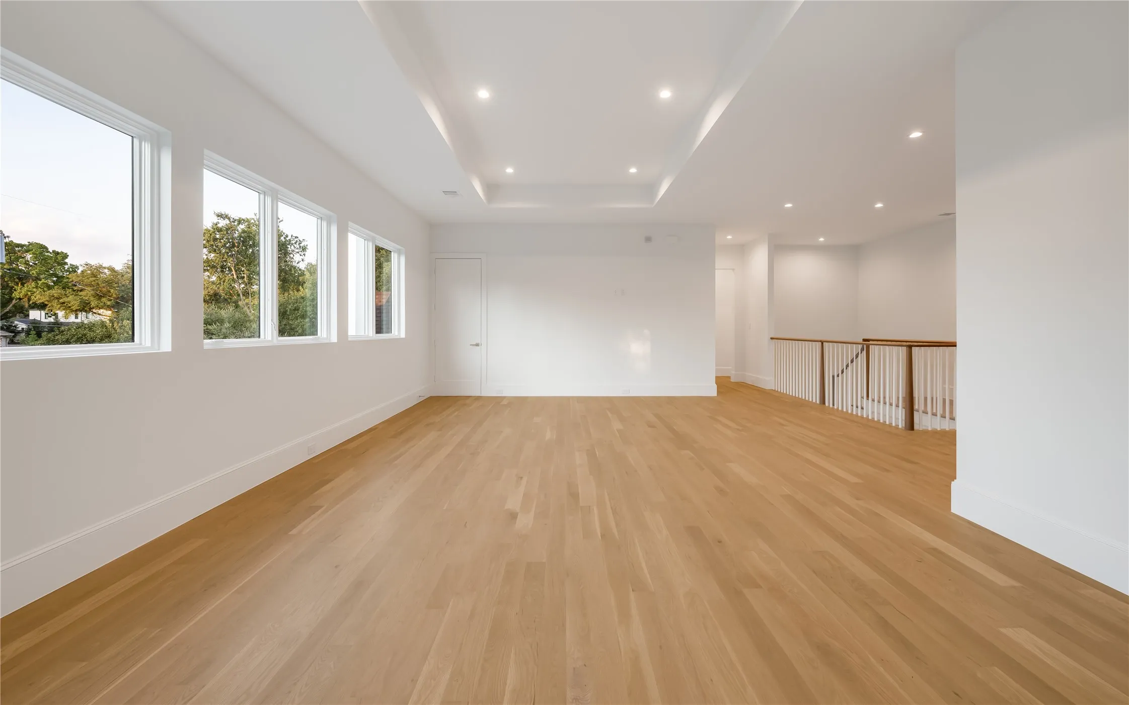 Expansive room featuring light wood-finish flooring, multiple windows, a tray ceiling with recessed lighting, and a white baluster railing