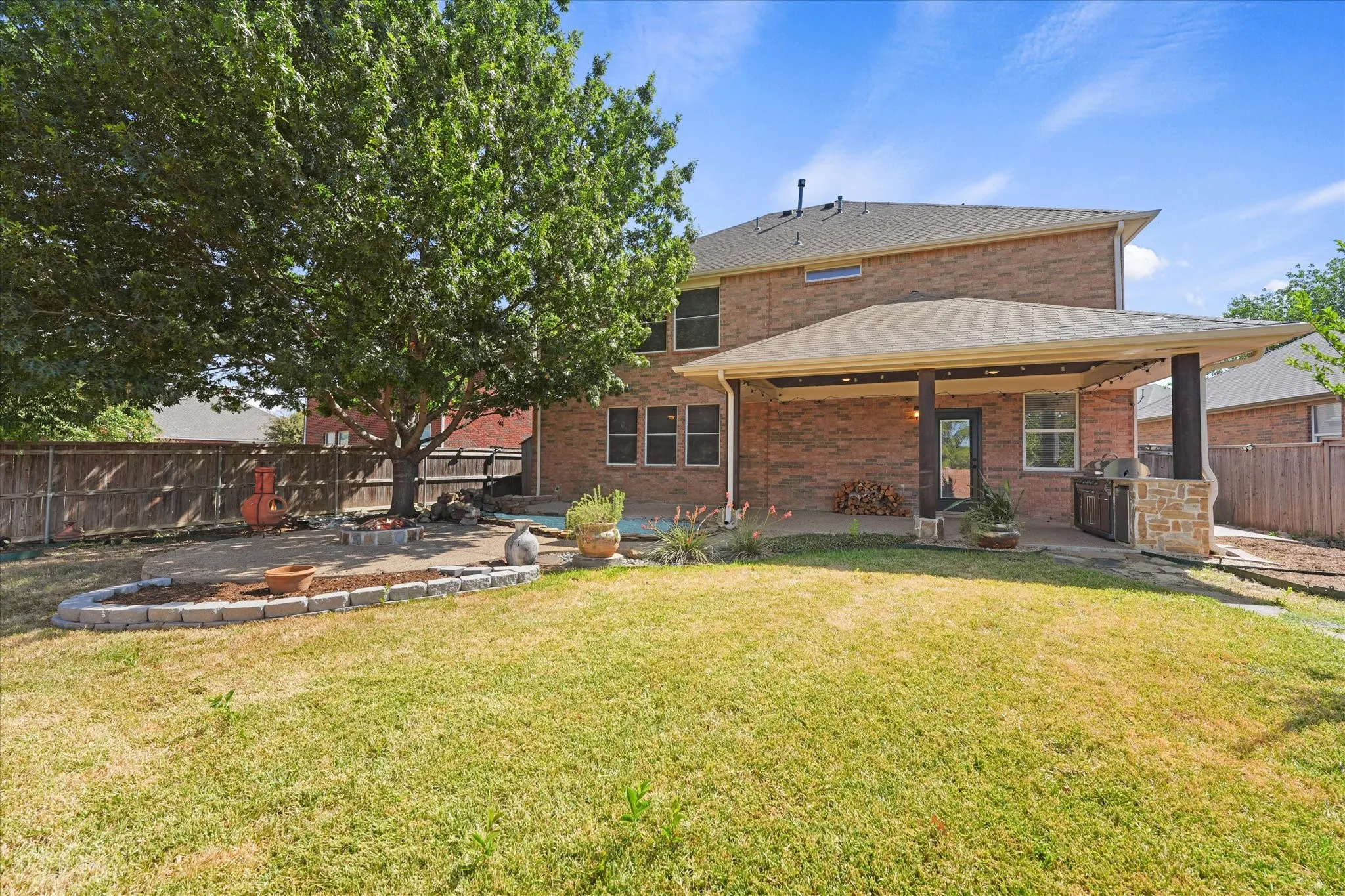 Expansive backyard featuring a covered patio, brick exterior, wood fence, and mature tree