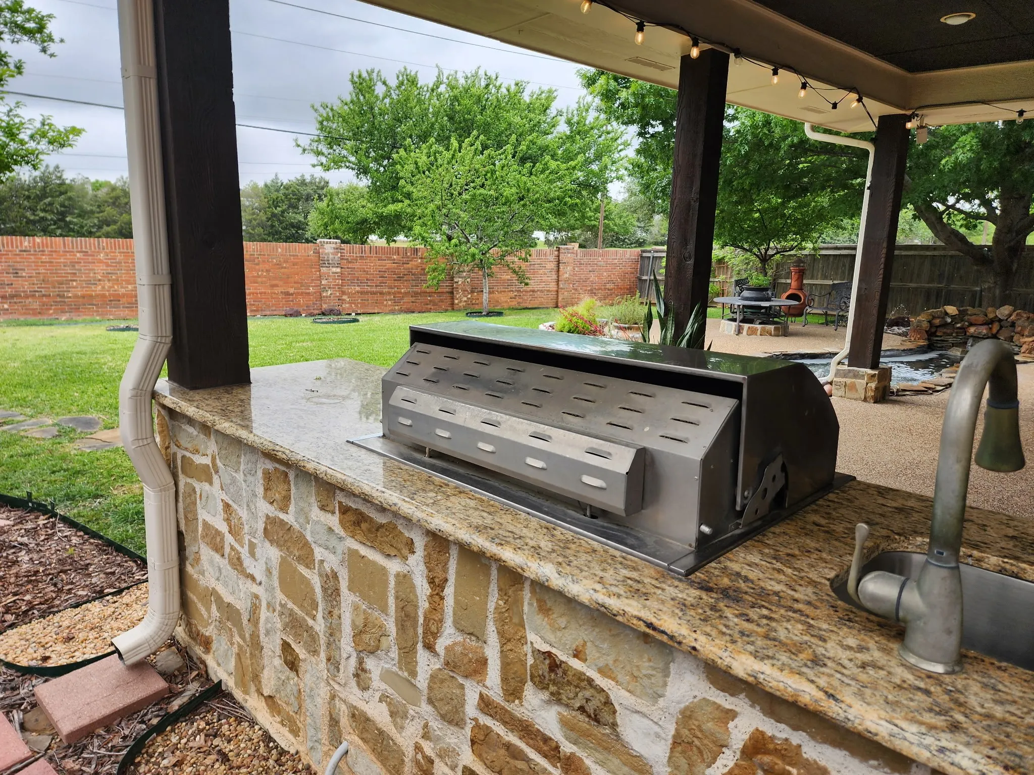 Outdoor kitchen featuring a built-in stainless steel grill, integrated sink with faucet, small refridgerator, stone veneer base, and granite countertop, all under a covered patio