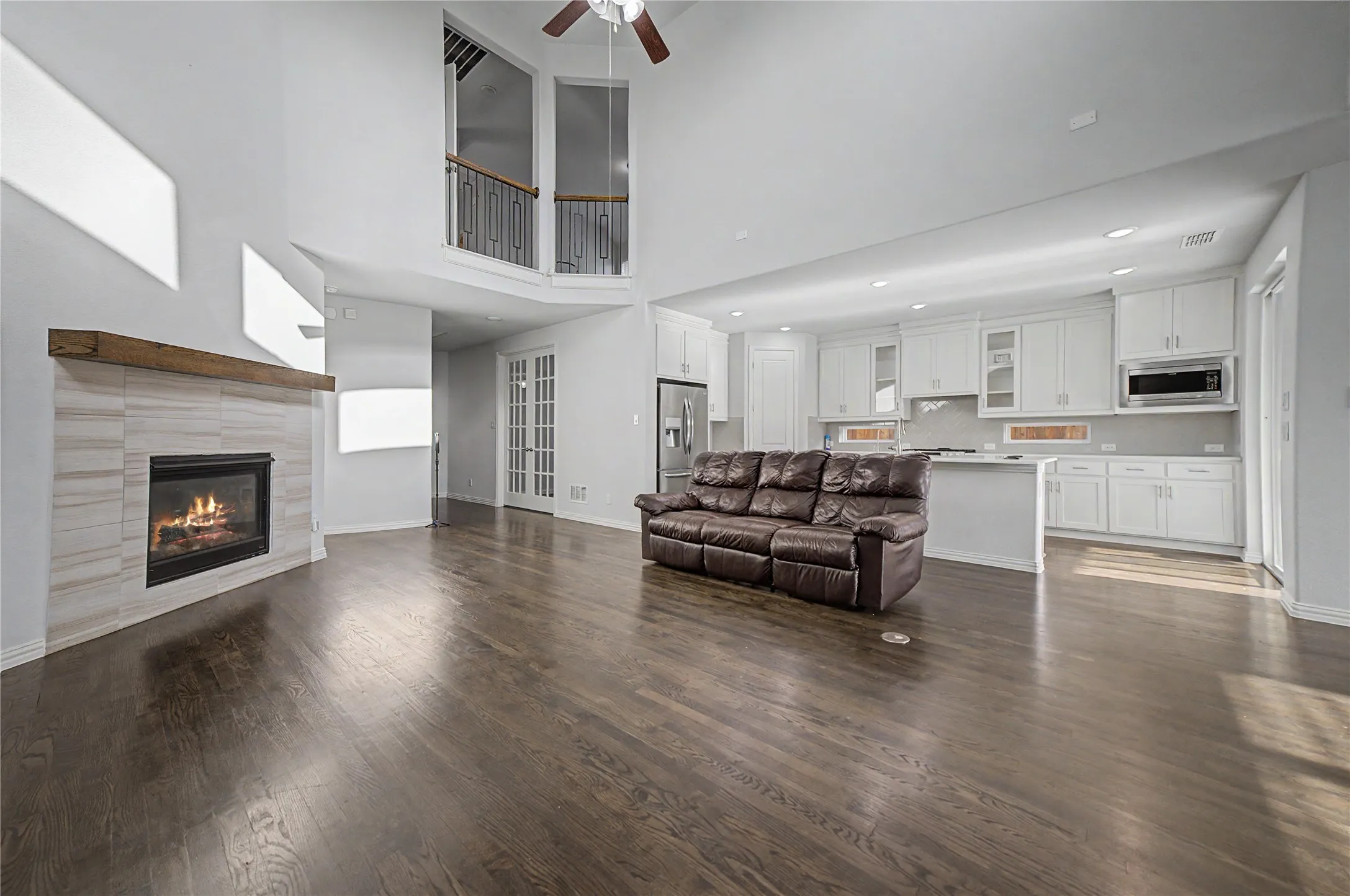 Hardwood-finish flooring throughout, two-story ceiling, gas fireplace with tile surround and wood mantel, white kitchen cabinetry with built-in microwave, and stainless steel appliances