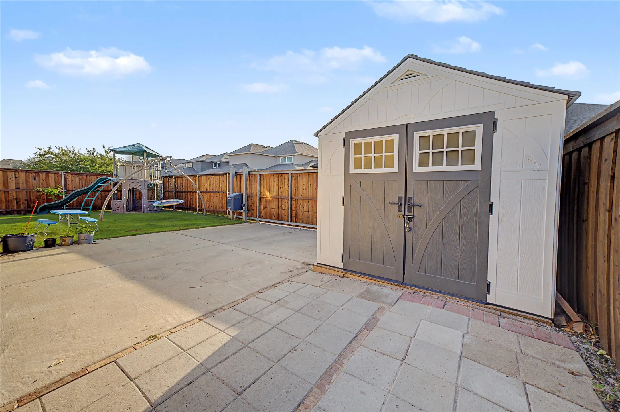 Spacious backyard retreat featuring a detached storage shed with dual gray doors and transom windows, expansive paved patio with geometric stone pavers, wooden privacy fencing, and manicured lawn space ideal for outdoor recreation and entertaining