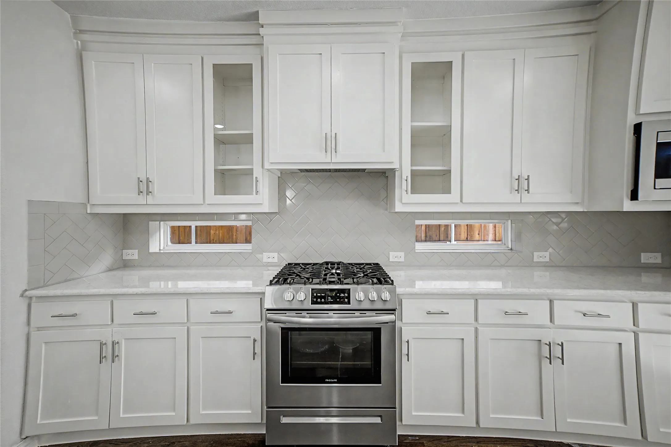 White shaker-style cabinetry, stainless steel range, light-toned countertops, herringbone pattern tile backsplash, and transom windows