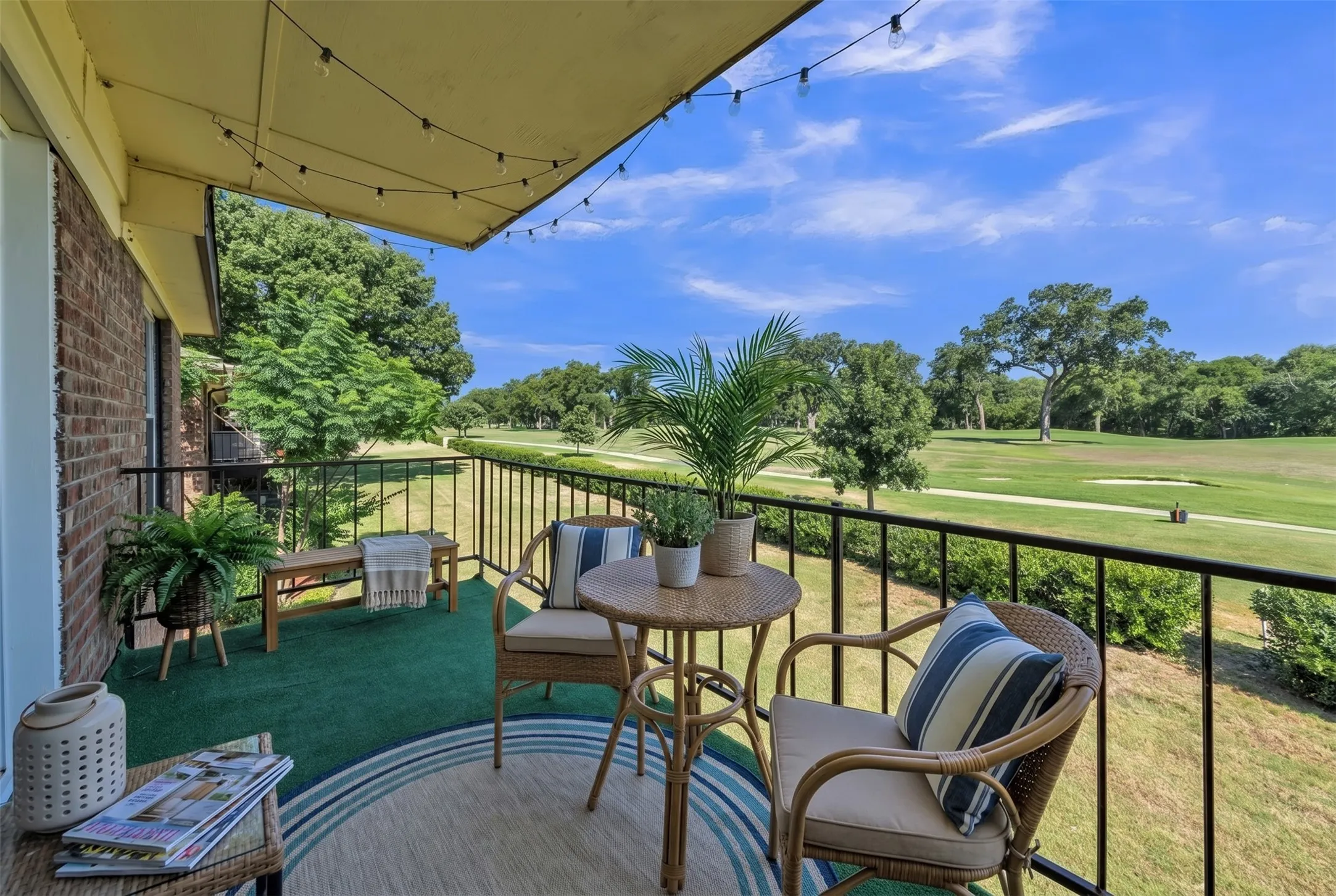 Balcony featuring a black wrought iron railing, brick exterior wall, overhead string lighting, and artificial turf flooring