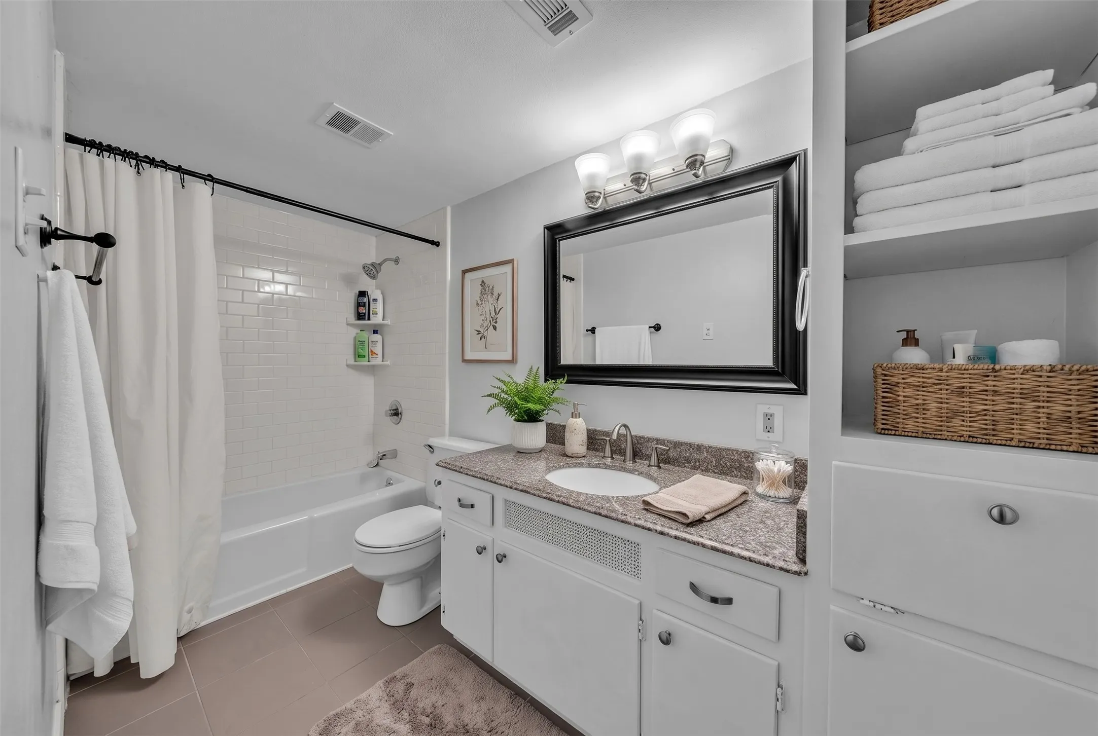 White subway tile shower surround, integrated white bathtub, built-in shelving, white vanity with stone-finish countertop, and recessed lighting