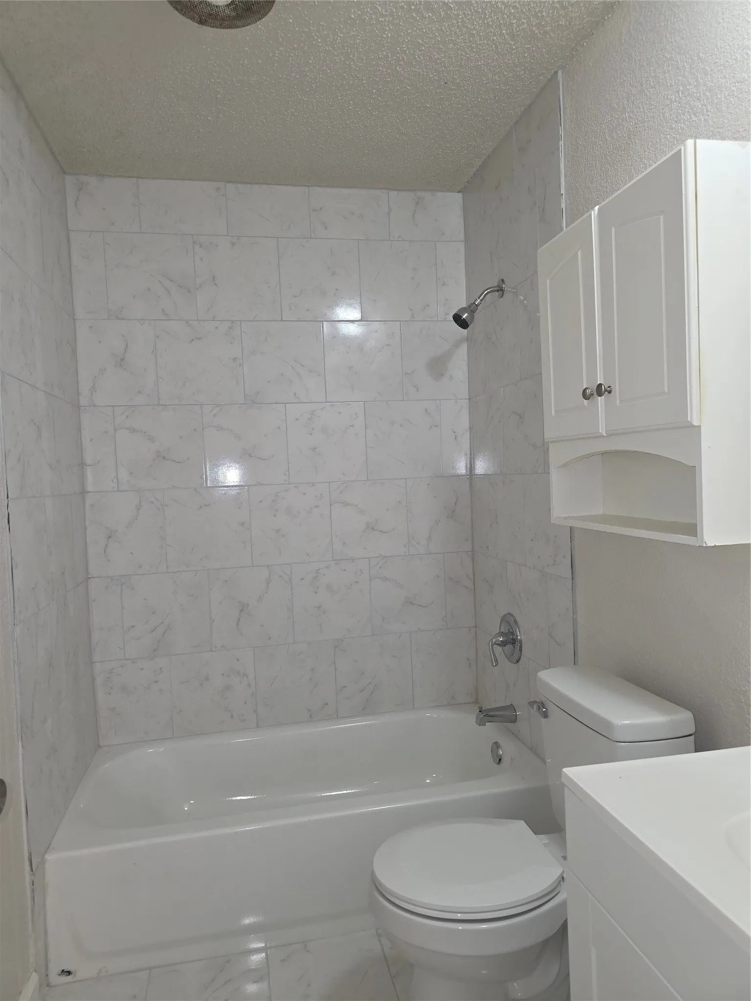 White subway tile shower surround with a built-in bathtub, a toilet, a chrome shower head, and a wall-mounted white storage cabinet