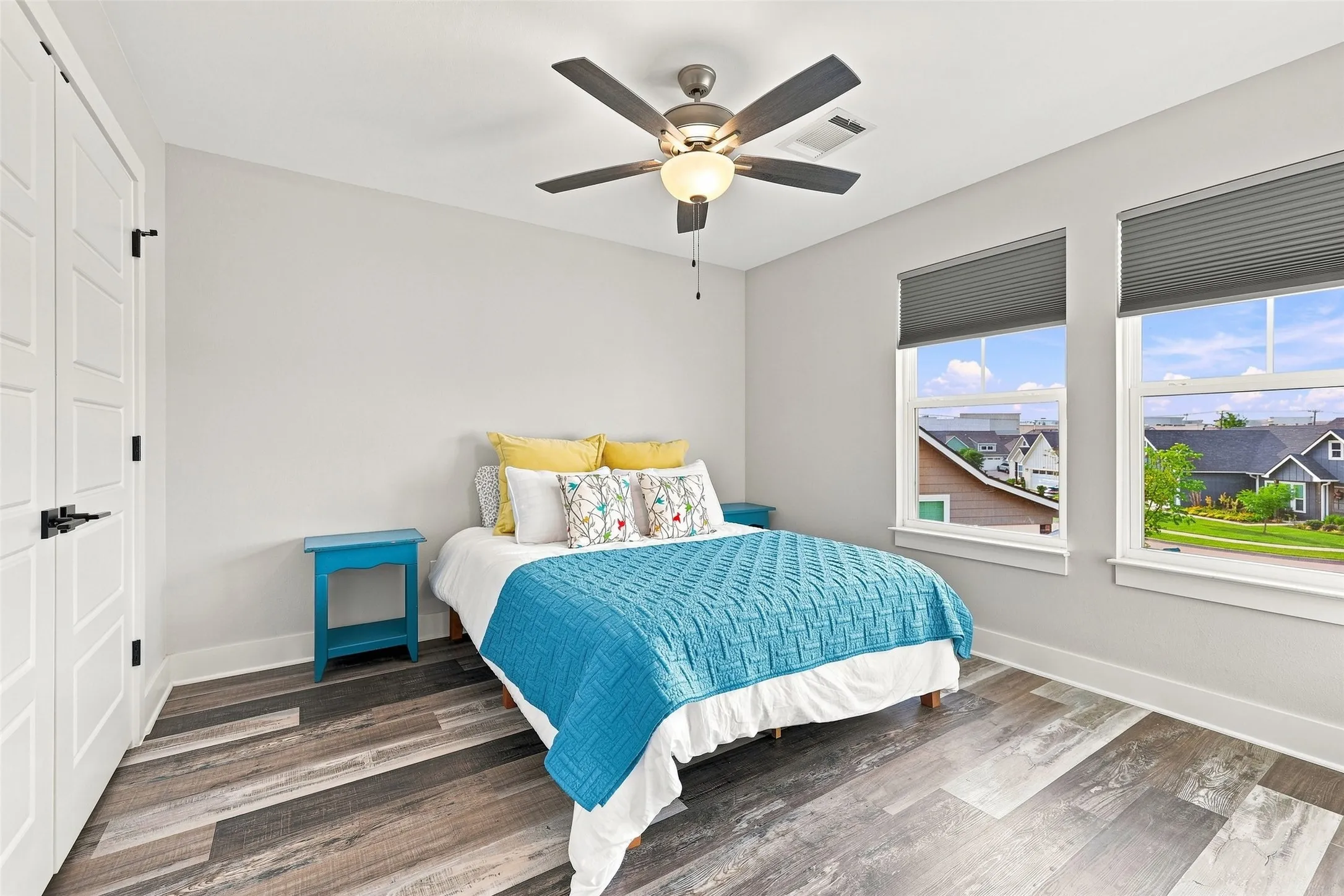 Wood-finish flooring, two large windows with roller shades, modern ceiling fan with integrated lighting, white paneled doors with black hardware, and light-toned wall paint