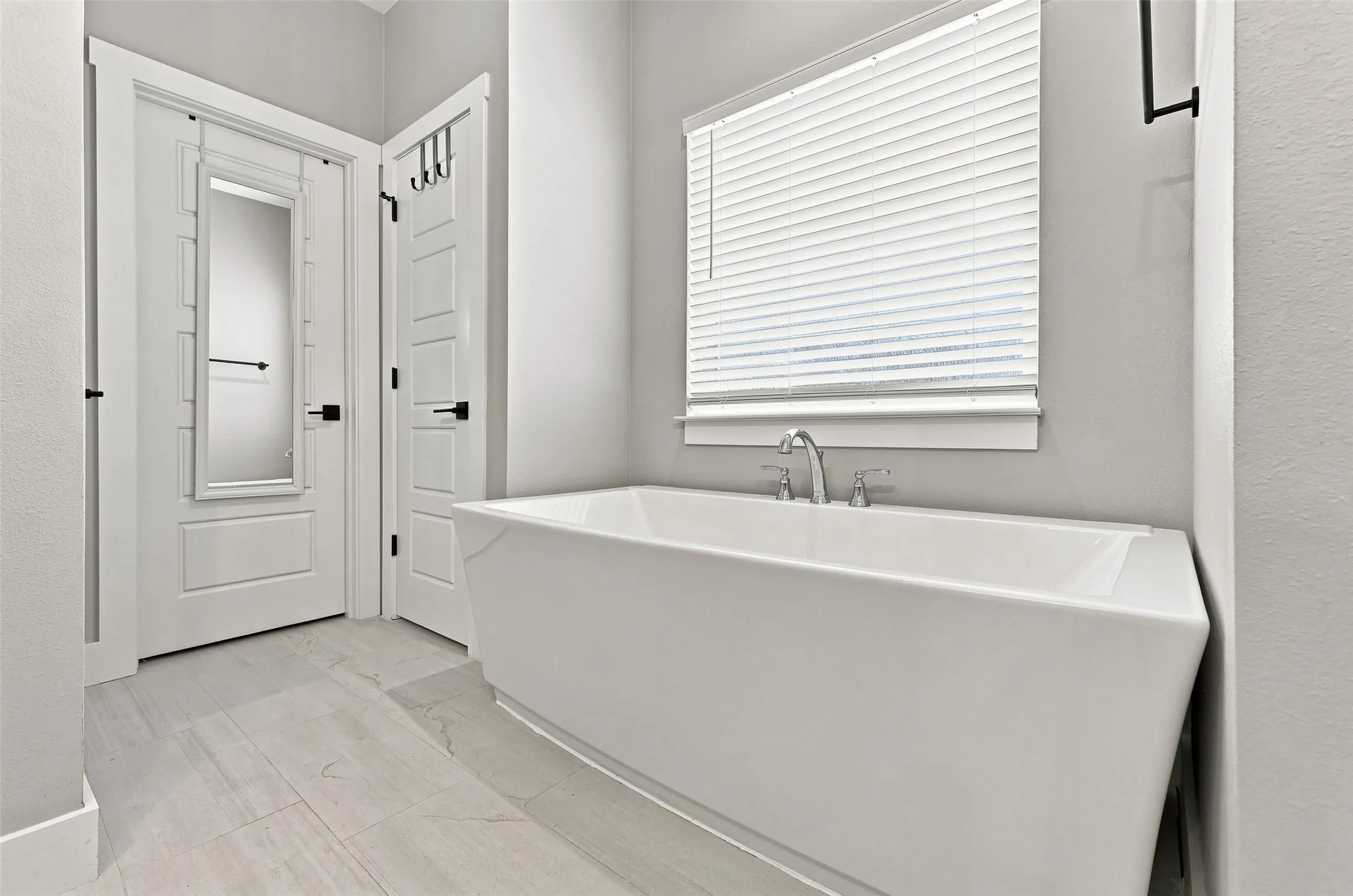 Freestanding tub with chrome fixtures, window with white blinds, light-toned tile flooring, and white paneled doors with black hardware