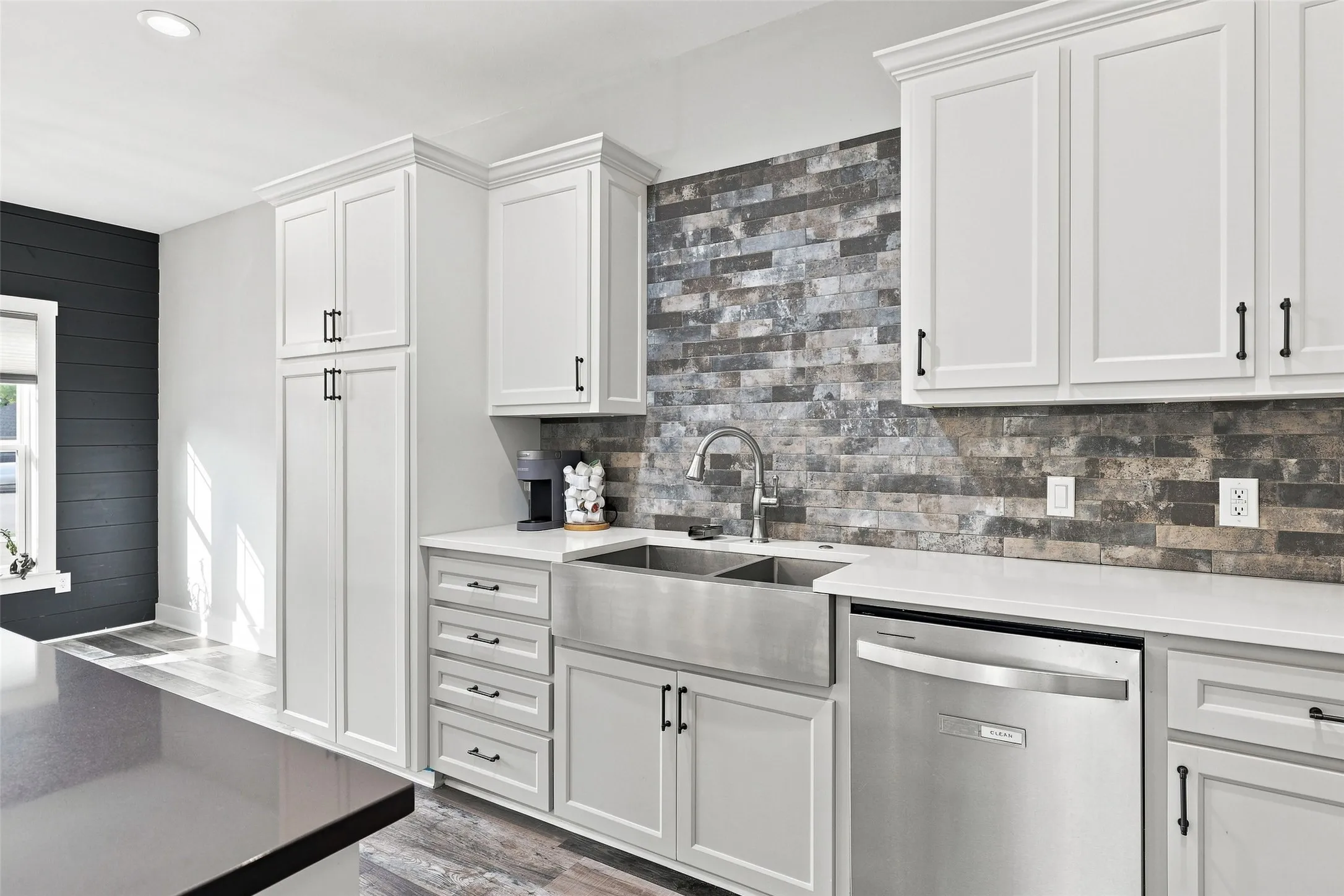 White shaker cabinetry, stone-look tile backsplash, stainless steel farmhouse sink, stainless steel dishwasher, and light-toned countertops