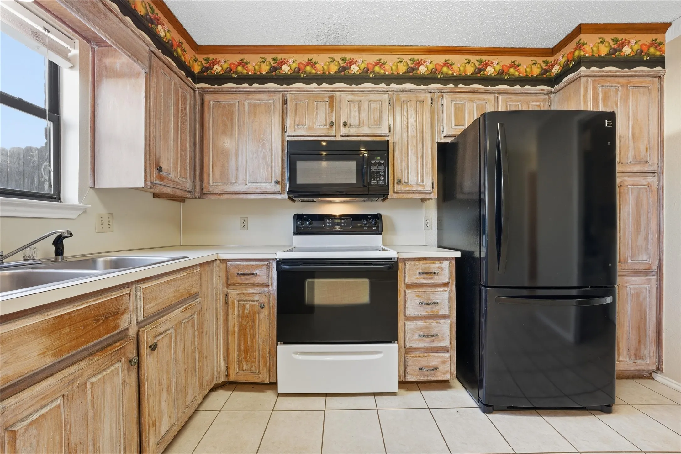 Kitchen featuring light wood-finish cabinetry, black and white appliances, a double basin sink, and tile flooring
