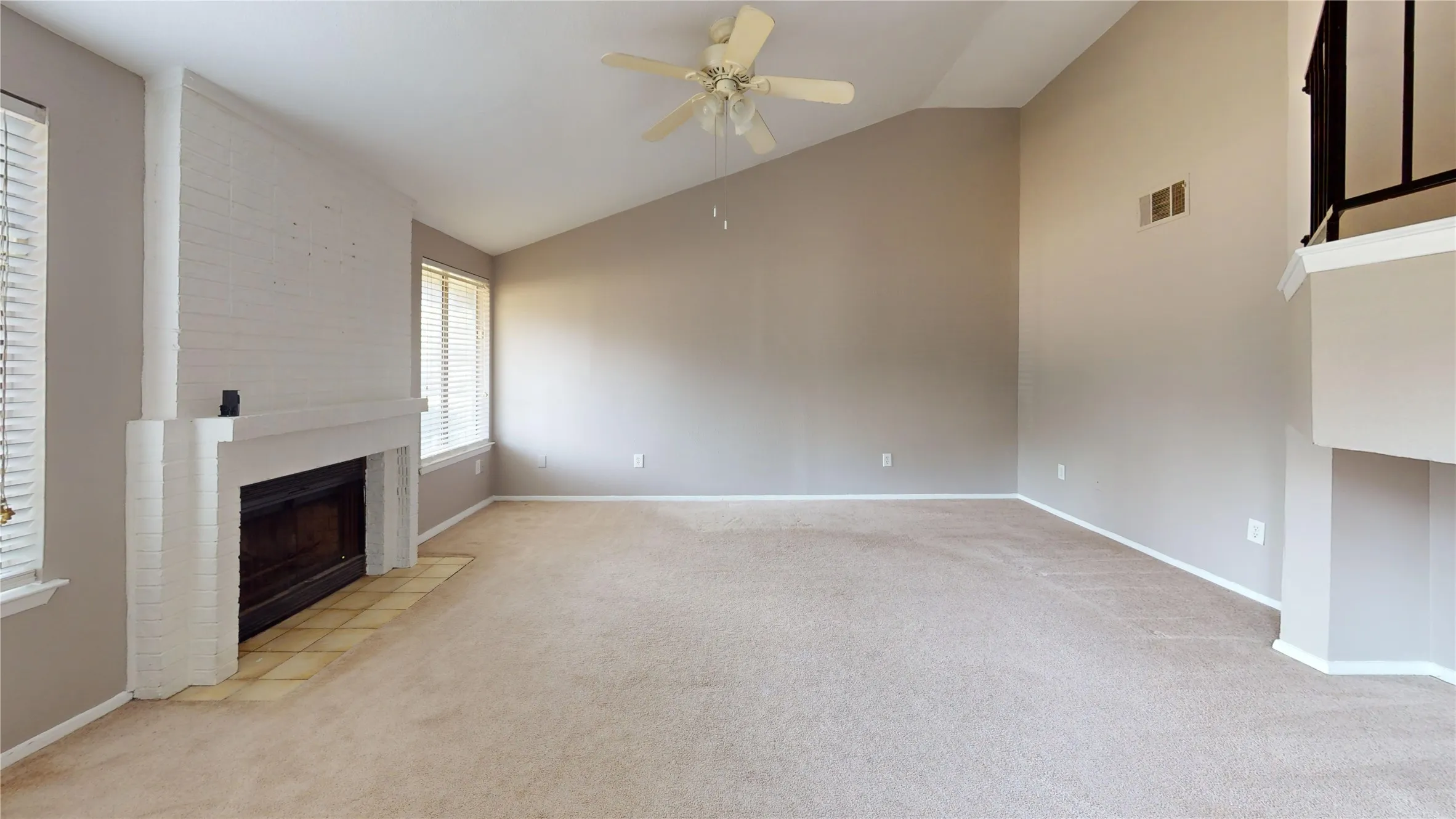 Carpeted living space featuring a white brick fireplace with hearth, vaulted ceiling, ceiling fan, and multiple windows