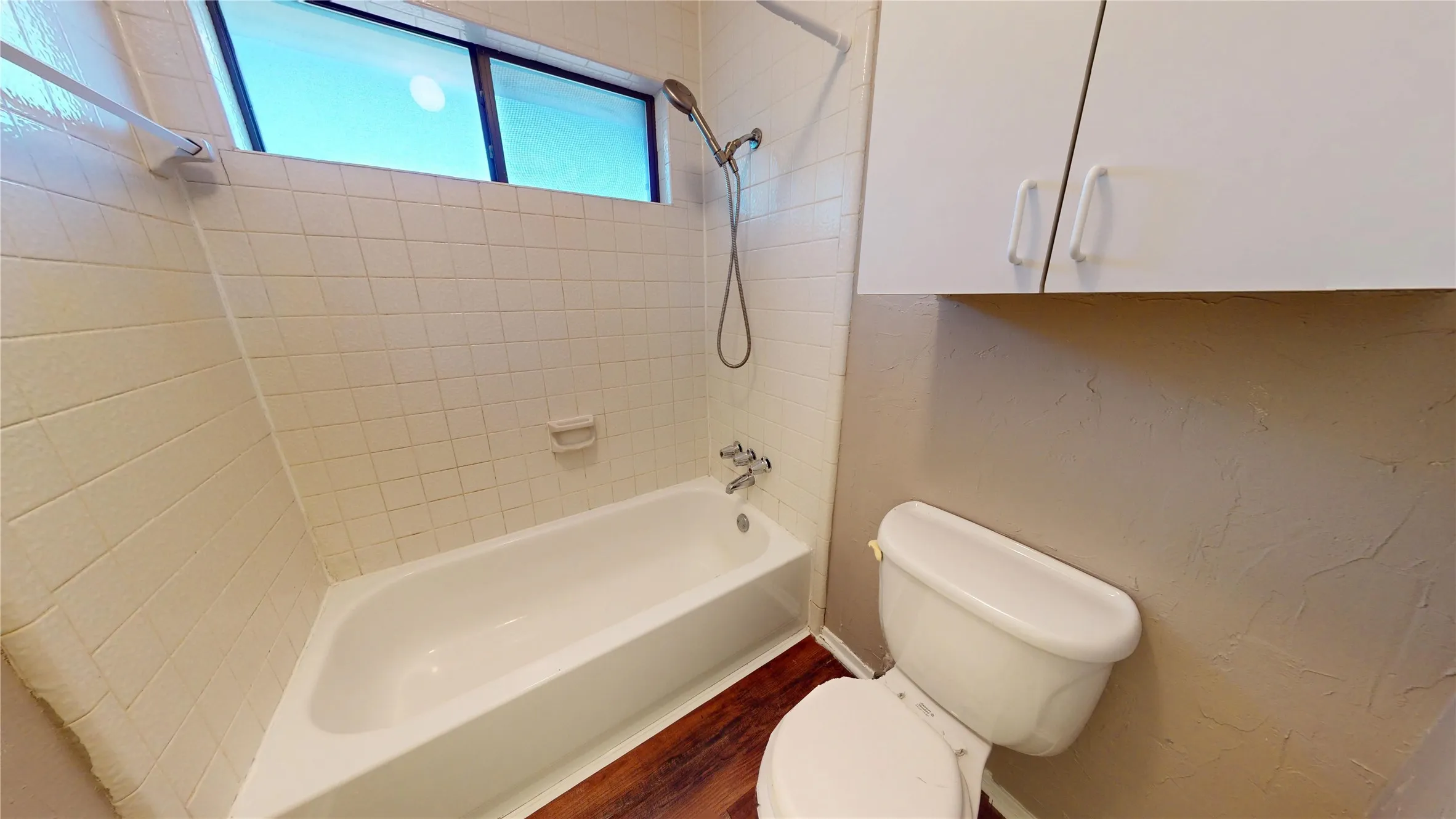 Bathtub with tile surround, wood-finish flooring, and upper cabinetry