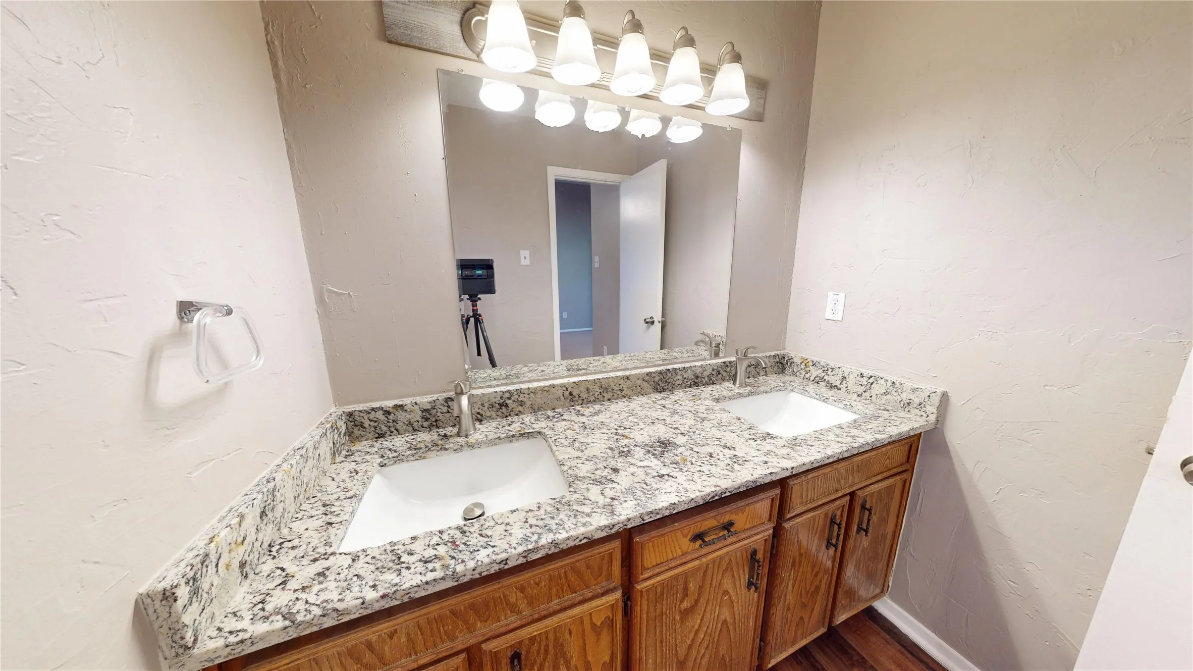 Dual undermount sinks with chrome faucets, granite-style countertop, wood-finish vanity cabinetry, and overhead vanity lighting
