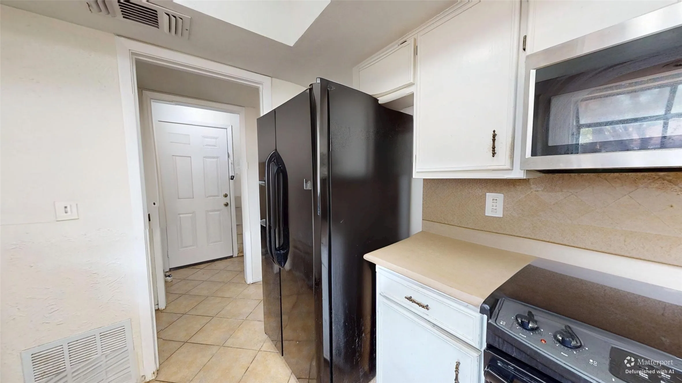 White paneled door, black side-by-side refrigerator, stainless steel microwave, white upper and lower cabinetry, and tiled flooring