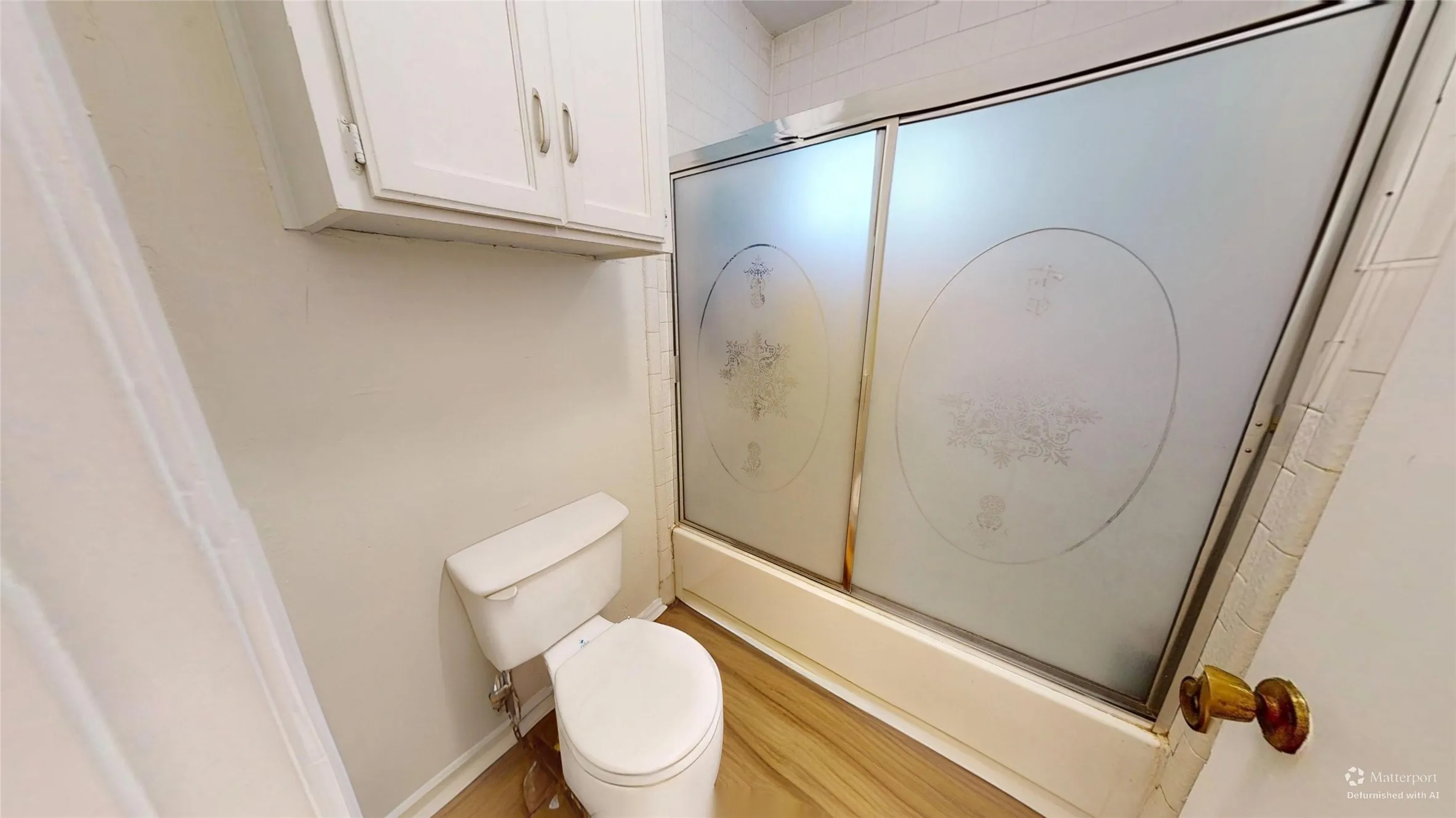 Built-in overhead white cabinetry, toilet, wood-finish flooring, and a shower with frosted sliding doors