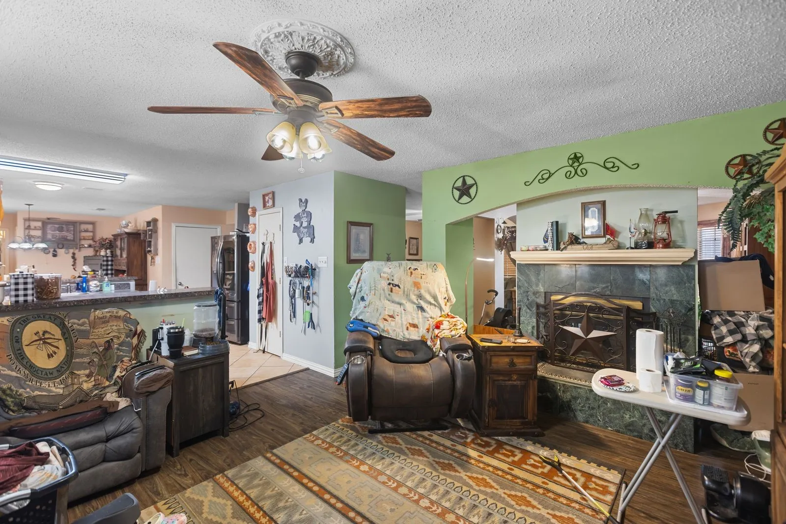 Wood-finish flooring throughout, ceiling fan with light fixture, decorative ceiling medallion, arched entryway, and a fireplace with a stone surround and mantel
