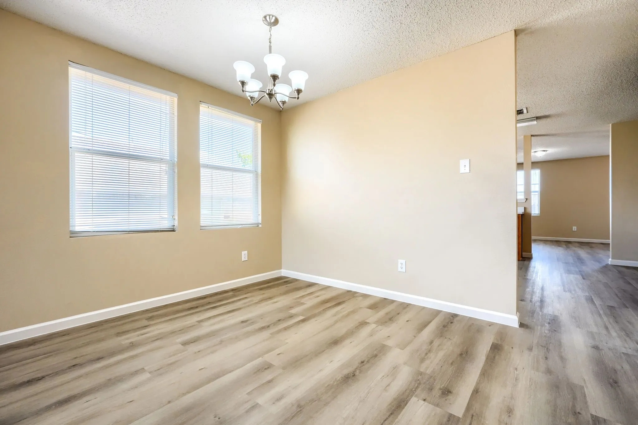 Wood-finish flooring throughout, two windows with blinds, neutral wall tones, and a brushed nickel chandelier with frosted glass shades
