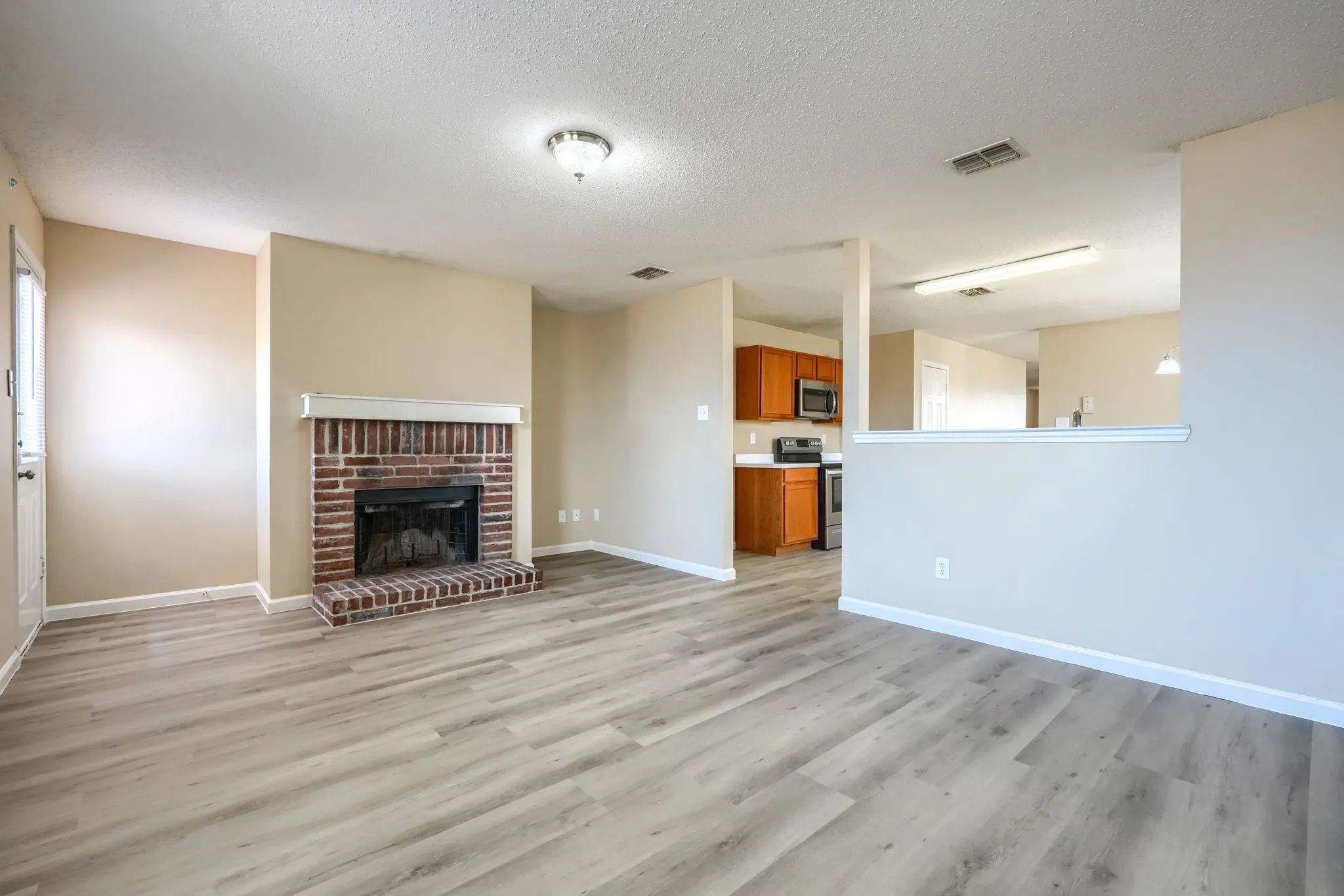 Brick fireplace with a white mantel, wood-finish flooring, recessed lighting, and kitchen visible