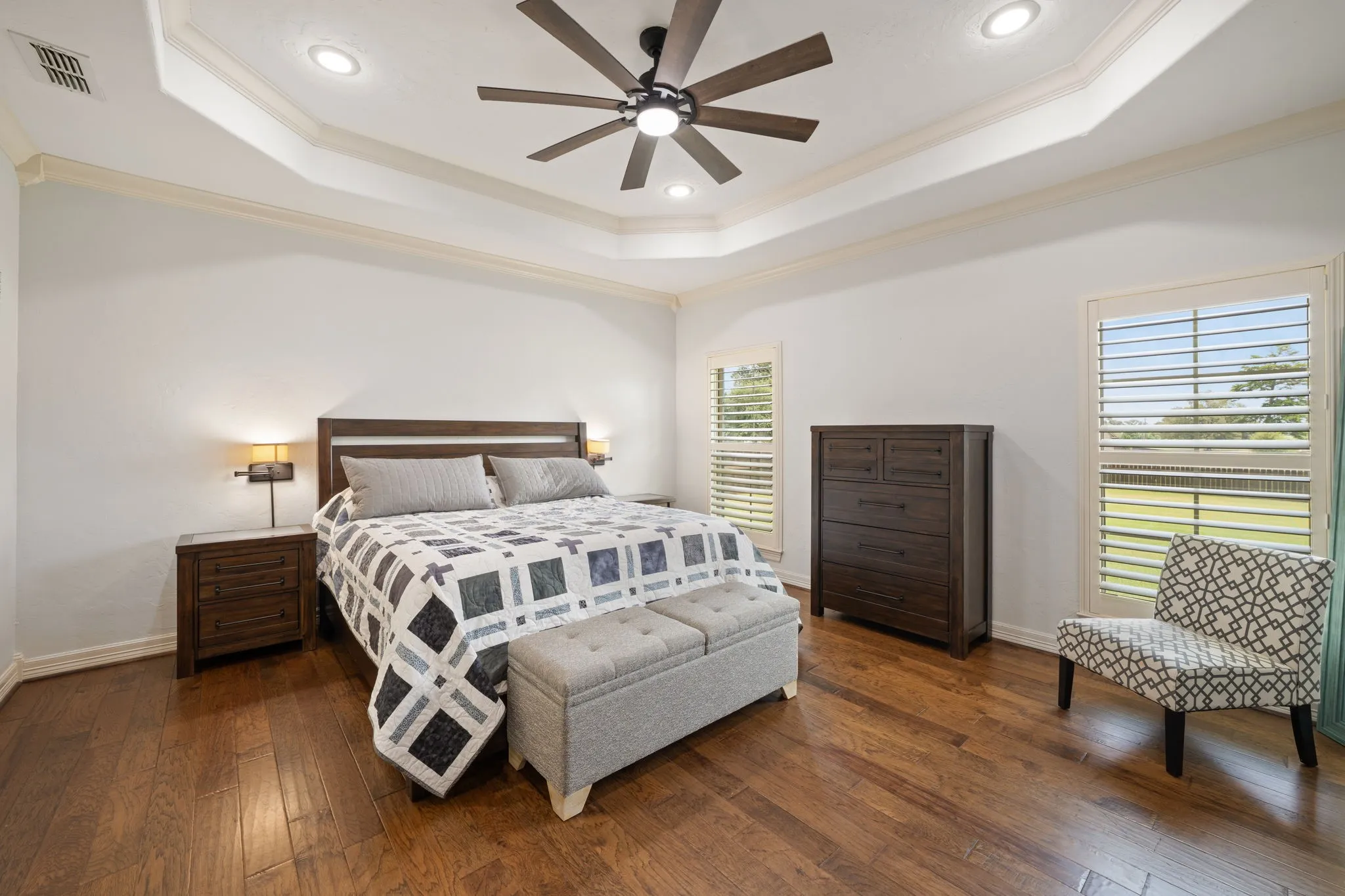 Tray ceiling with recessed lighting, wood-finish flooring, ceiling fan with integrated lighting, window shutters, and multiple windows