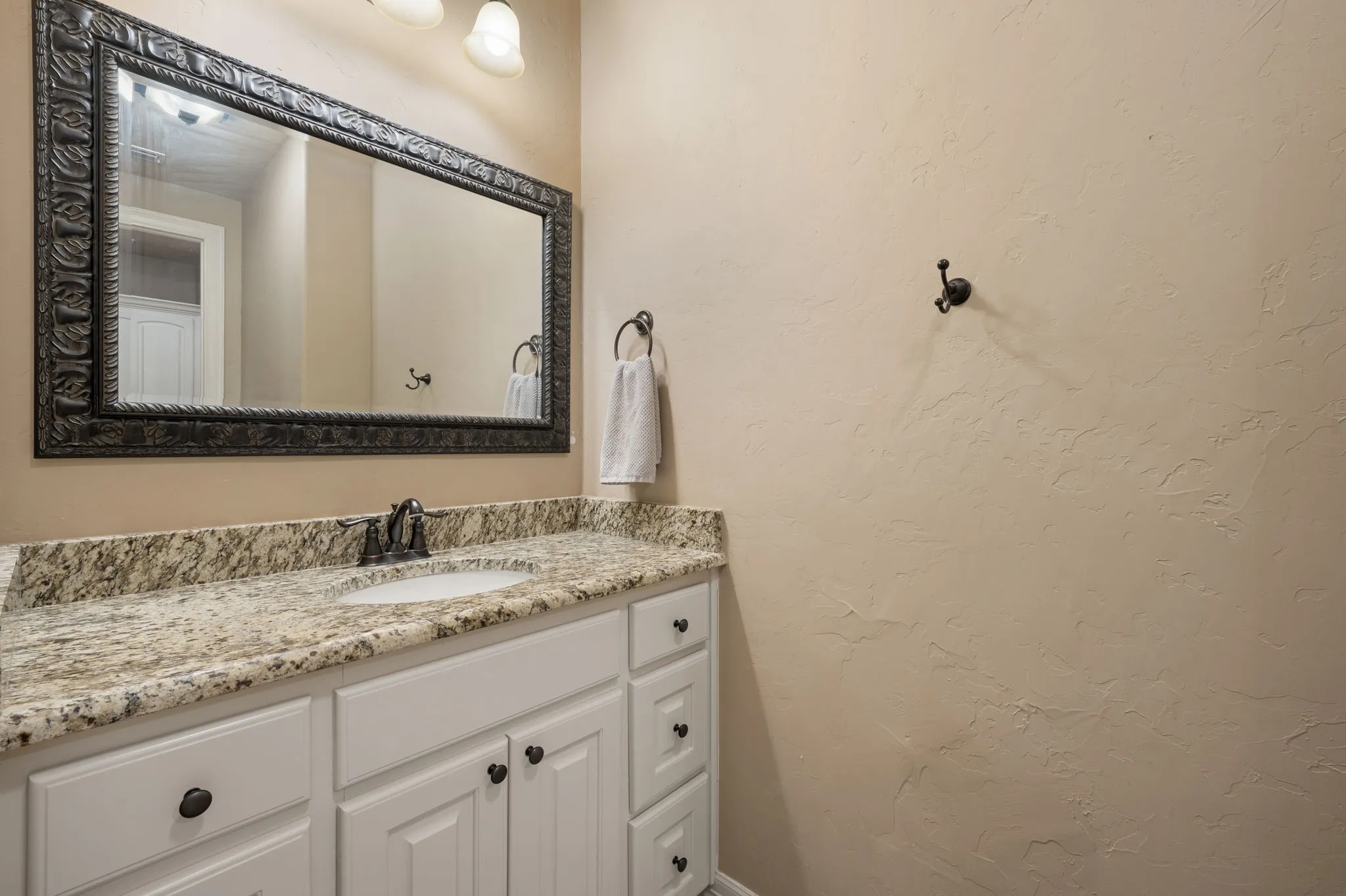 Elegant bathroom vanity featuring white cabinetry with oil-rubbed bronze hardware, a granite-look countertop with undermount sink, and a decorative framed mirror with ornate detailing
