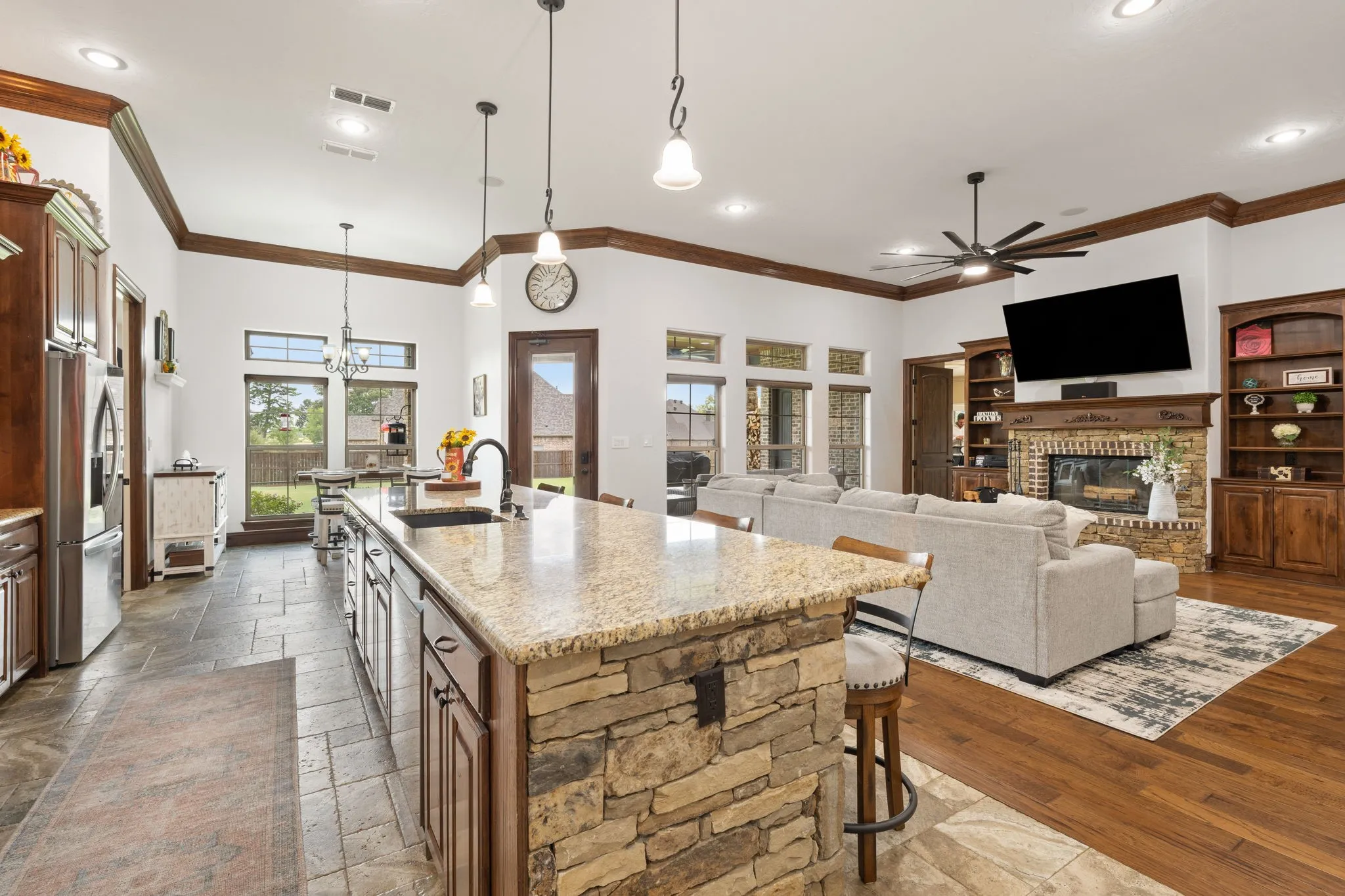 Open-concept kitchen and living area featuring a large center island with stone base and granite countertop, stainless steel appliances, white cabinetry with glass-front upper cabinets, pendant lighting, ceiling fan, recessed lighting, hardwood flooring, brick fireplace with built-in shelving, and expansive windows overlooking the landscape