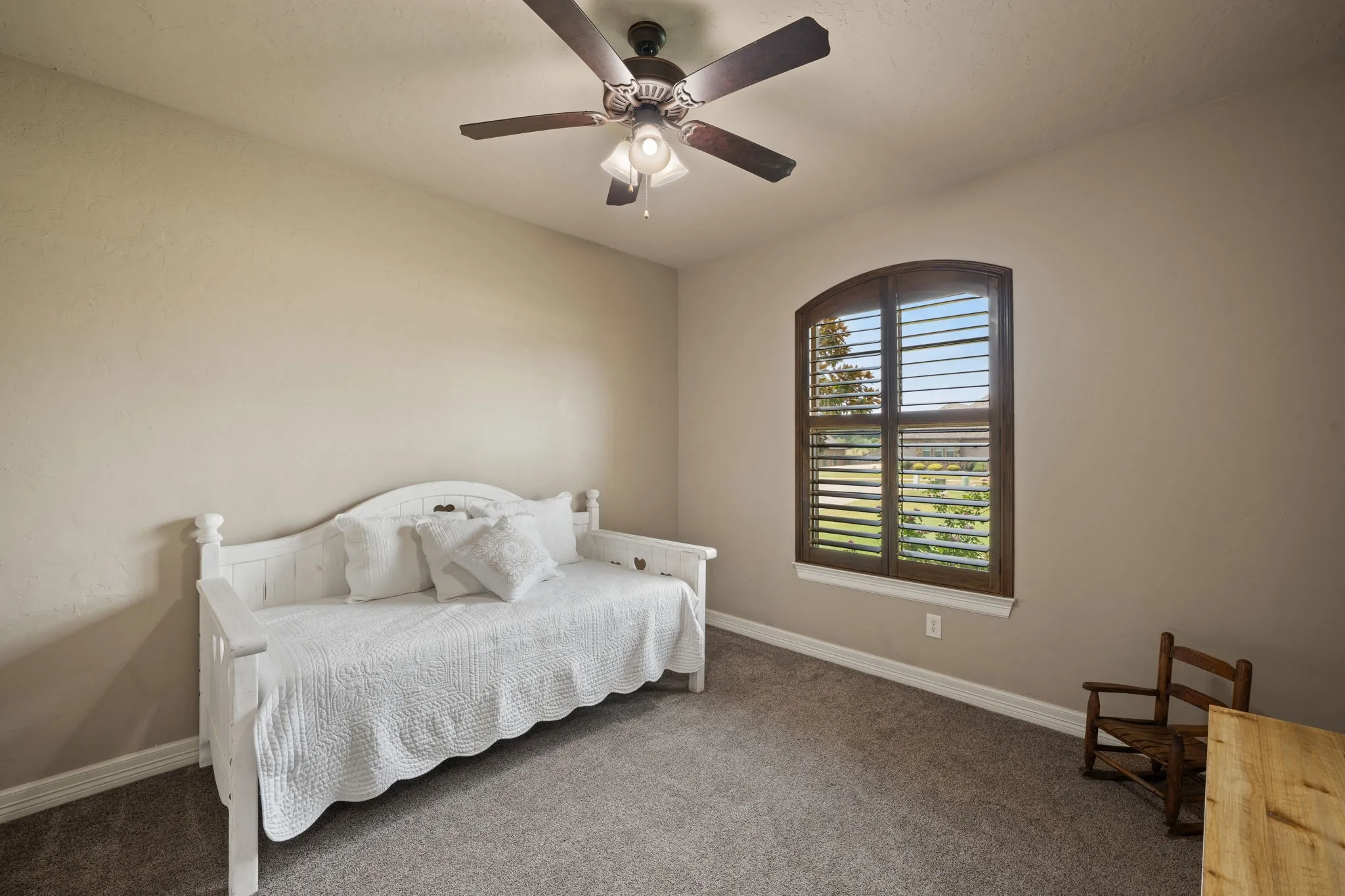 Bright secondary bedroom featuring neutral carpeting, a ceiling fan with integrated lighting, and an arched window with plantation shutters offering natural illumination and outdoor visibility