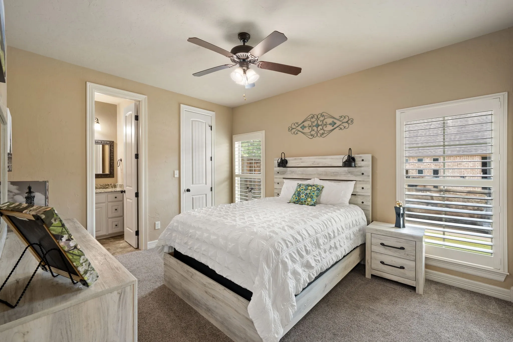 Master bedroom featuring a ceiling fan with integrated lighting, wood-finish bed frame, nightstand with drawer storage, white painted door frames, plantation shutters on glass doors, and neutral wall-to-wall carpeting throughout