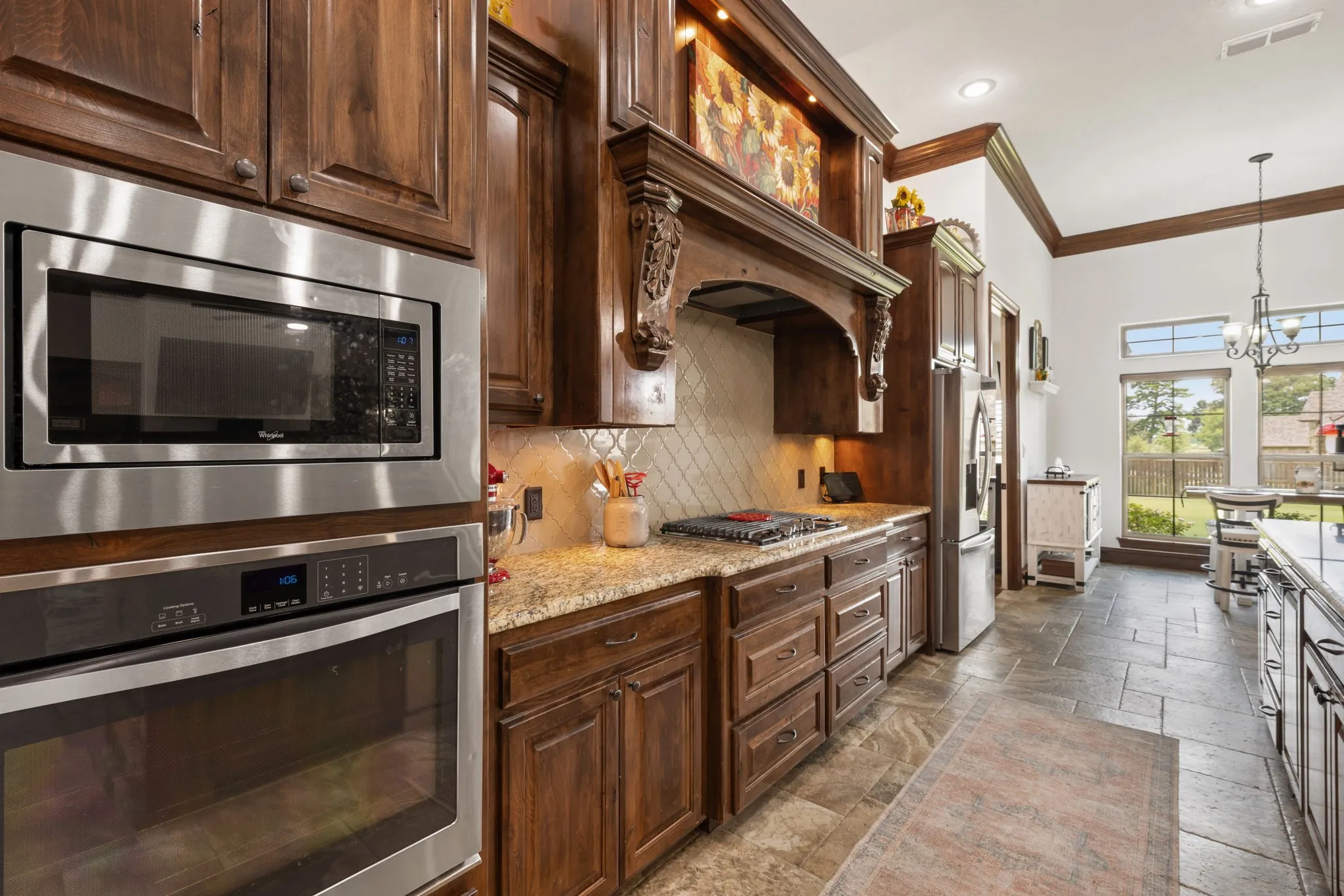 Stainless steel built-in microwave and oven, wood cabinetry with decorative range hood, light-toned granite countertops, tiled backsplash, and stone tile flooring
