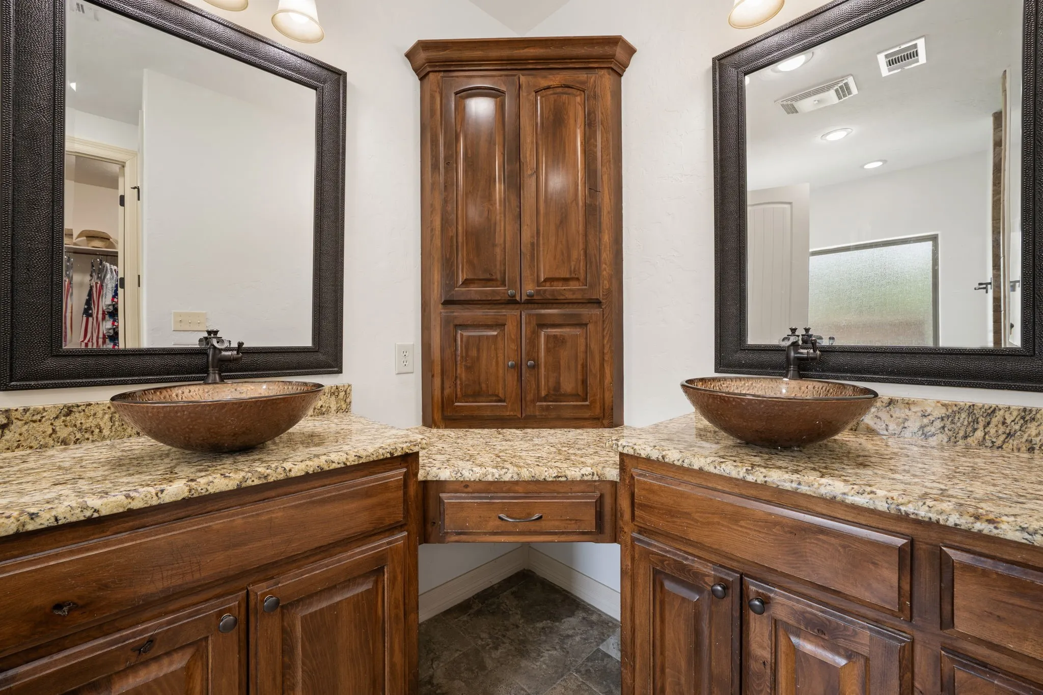 Dual vanity bathroom featuring paired vessel sinks with polished stone countertops, wood cabinetry with bronze hardware, framed mirrors with dark trim, and a central wood-finish cabinet with four-panel doors