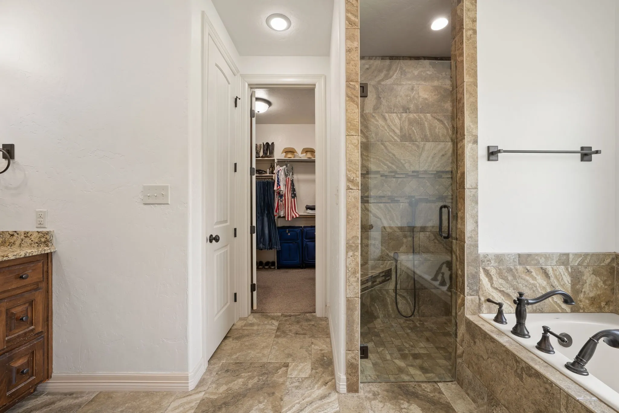 Walk-in shower with clear glass enclosure and stone-look tile surround, built-in soaking tub with dark bronze fixtures, wood-finish vanity with stone-finish countertop, and stone-look tile flooring