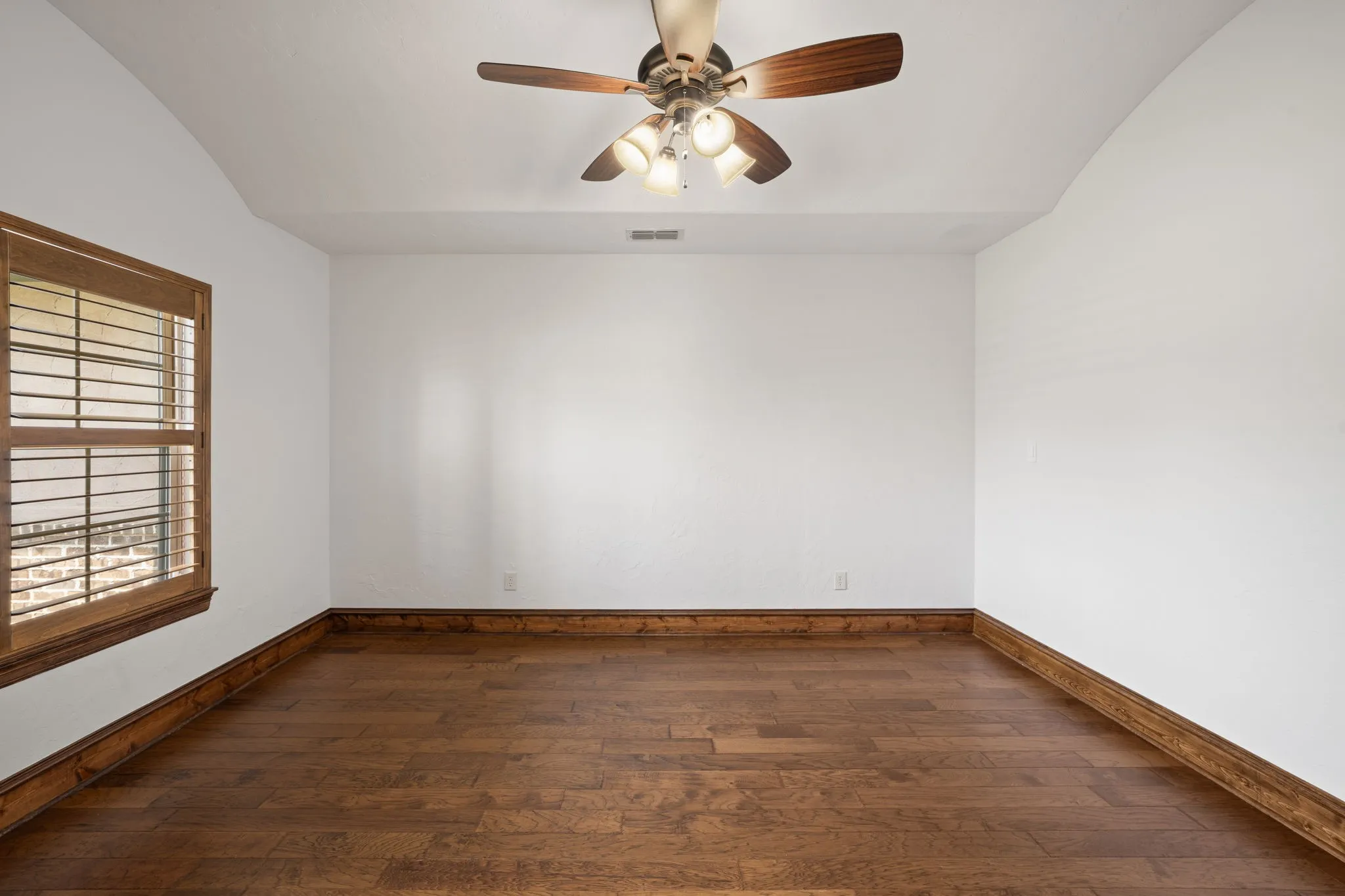 Spacious room featuring rich wood-finish flooring with coordinating trim detail, white walls, ceiling fan with integrated lighting, and a shuttered window allowing natural light