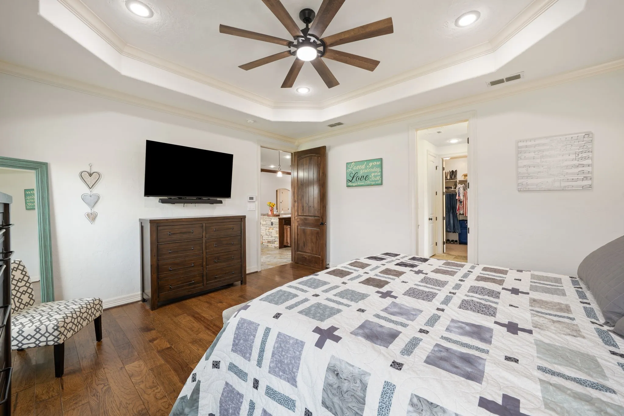 Spacious primary bedroom featuring a tray ceiling with recessed lighting and wood-blade ceiling fan, wall-mounted flat-screen television above dark wood cabinetry, hardwood flooring throughout, and multiple windows providing natural light