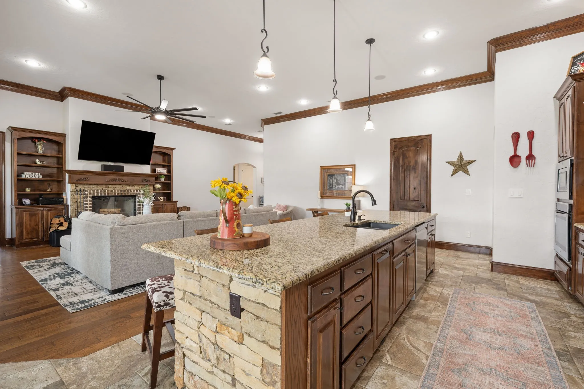Open-concept kitchen and living area featuring a substantial island with stone-clad base and wood cabinetry, granite countertops, stainless steel appliances, and pendant lighting