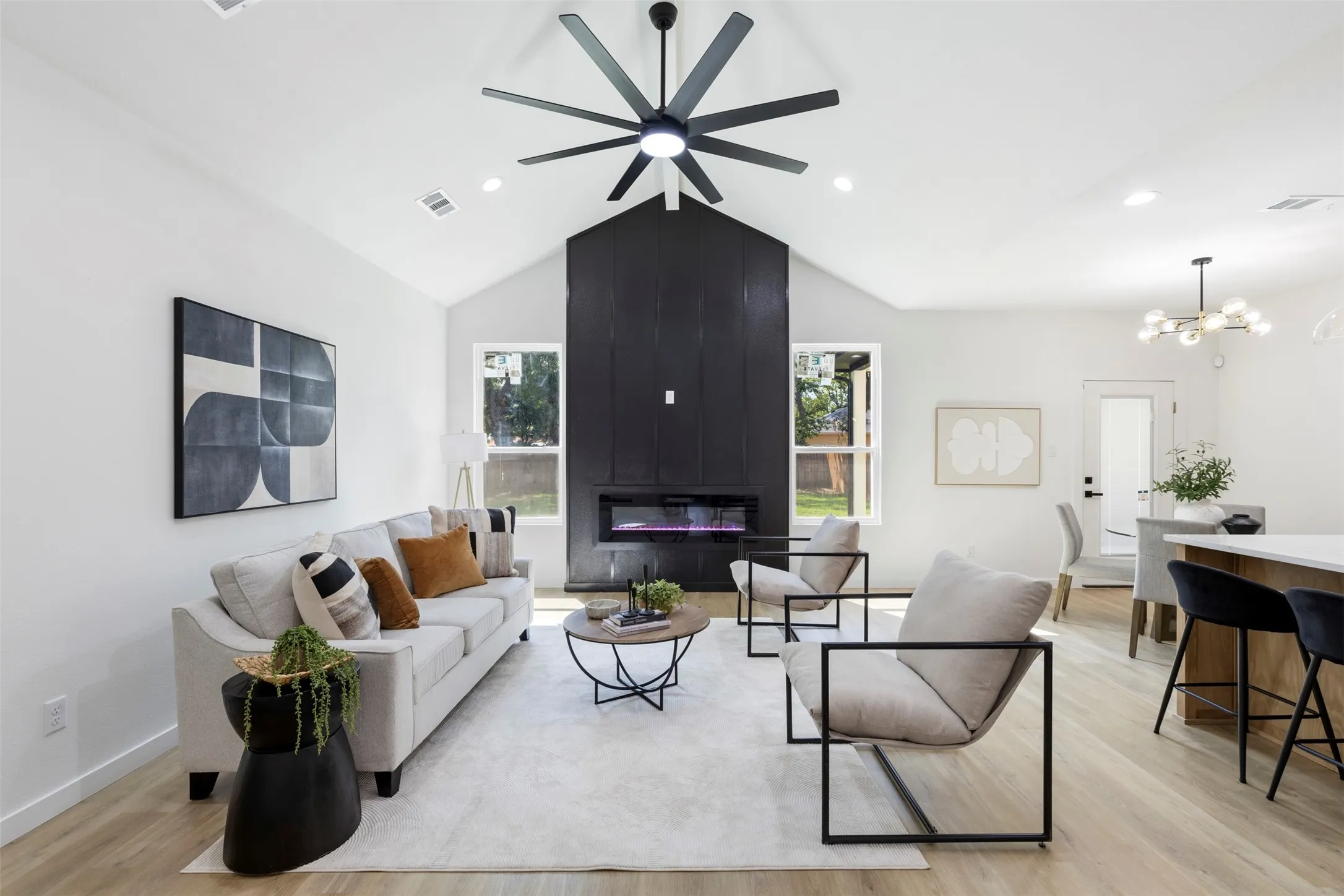 Open-concept living space anchored by a striking black fireplace chimney with modern ceiling fan, wood-finish flooring throughout, and abundant natural light from multiple windows
