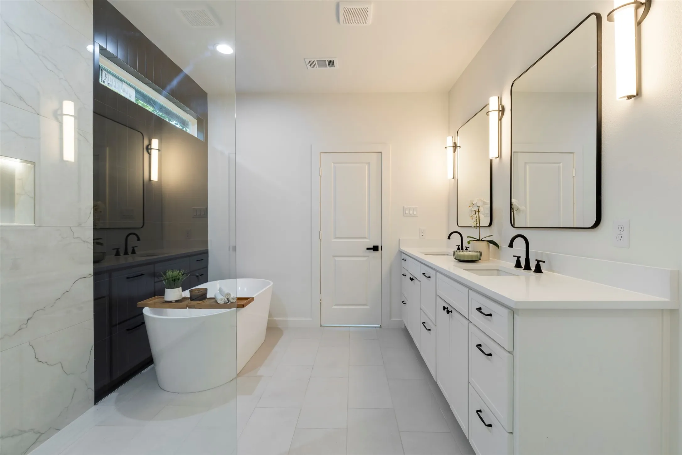Dual vanities with white countertops, rectangular mirrors with integrated lighting, a freestanding soaking tub, and a glass-enclosed shower with large format tile