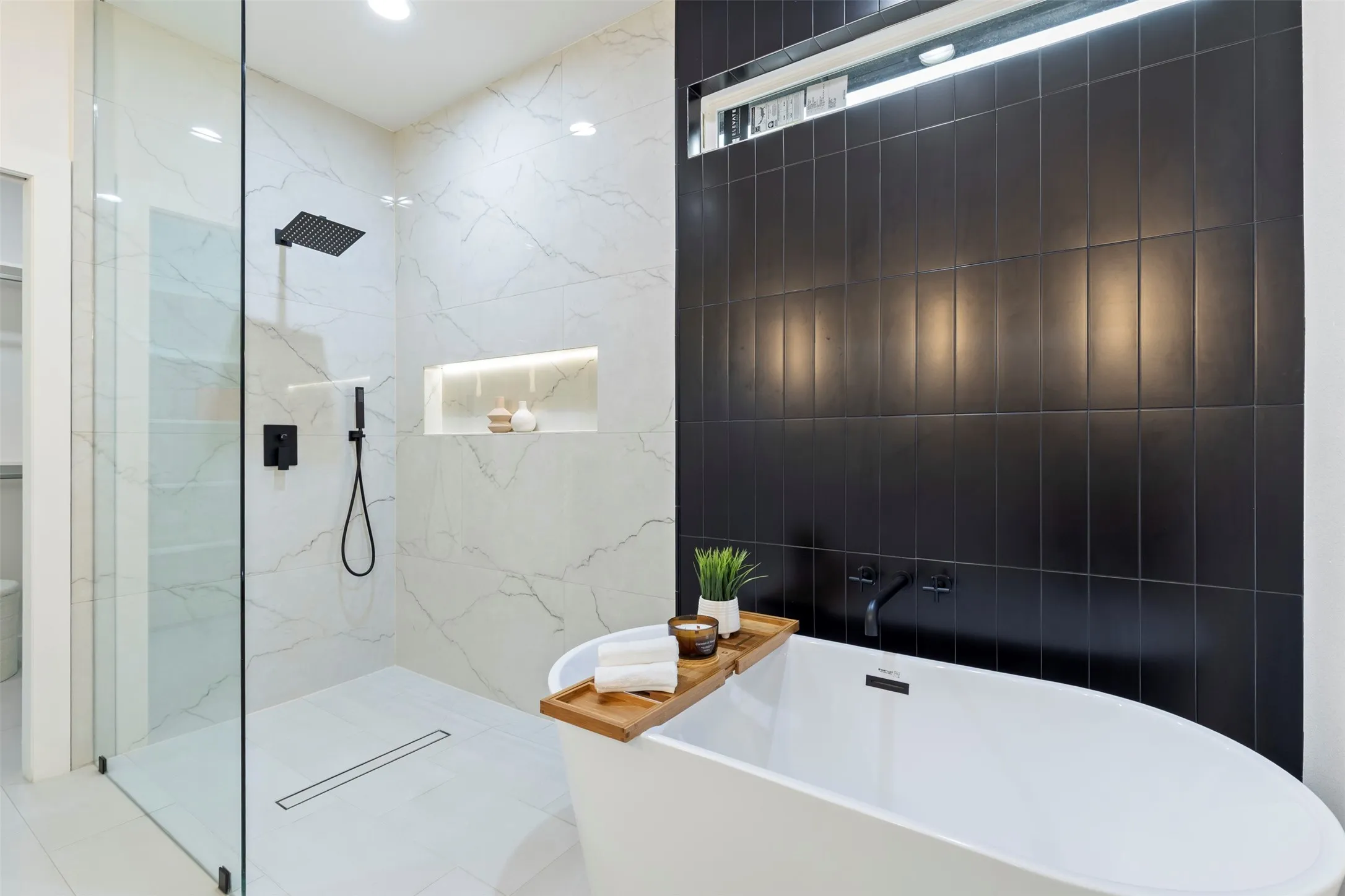 Contemporary primary bathroom featuring a freestanding soaking tub with integrated deck space, a walk-in shower with frameless glass enclosure and rainfall showerhead, matte black subway tile accent wall with integrated lighting, white tile flooring, recessed ceiling lighting, and a built-in niche with LED accent lighting