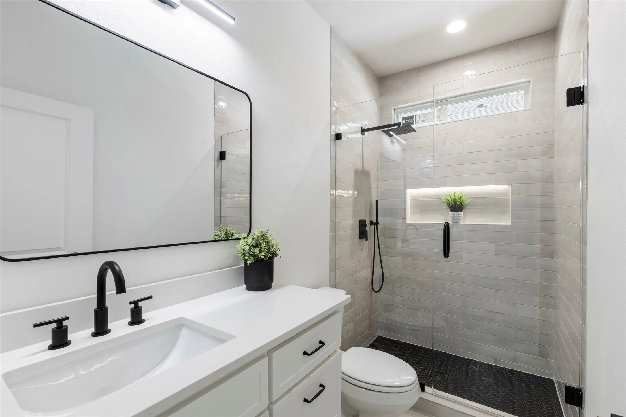 Vanity with integrated sink and matte black hardware, framed rectangular mirror, glass-enclosed shower, tiled shower walls, and a recessed shower niche with integrated lighting