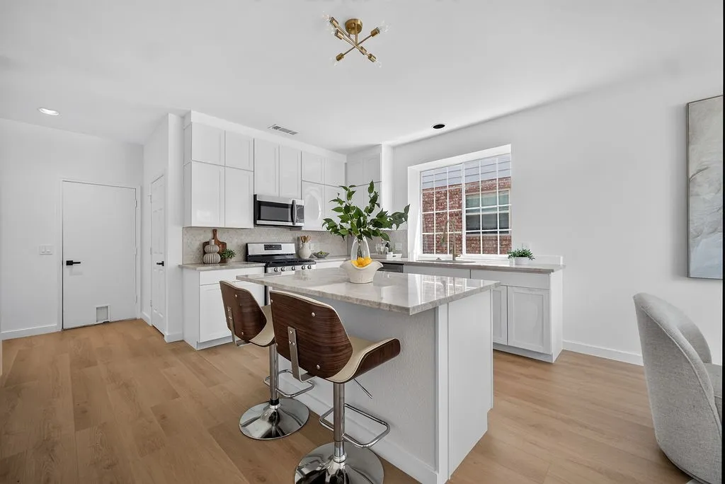 Bright, open-concept kitchen featuring white cabinetry, stainless steel appliances, a large quartz-topped island with seating for two, and abundant natural light from multi-pane windows
