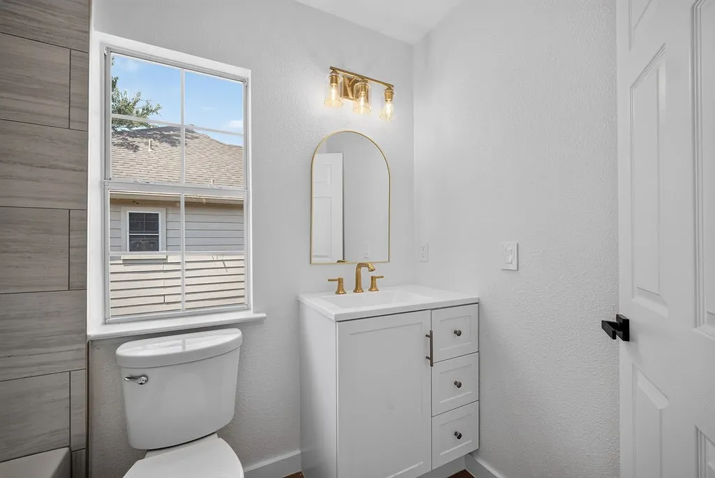 Bright, updated bathroom featuring a white vanity with brushed gold hardware, arched mirror, quartz countertop, and natural light from a large window