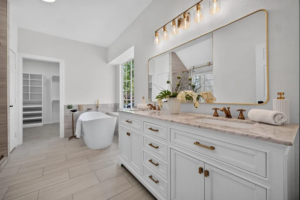 Spa-inspired primary bathroom featuring a freestanding soaking tub, dual-sink vanity with marble-finish countertops, brass hardware and fixtures, large framed mirror with integrated lighting, and wide-plank wood-finish flooring throughout