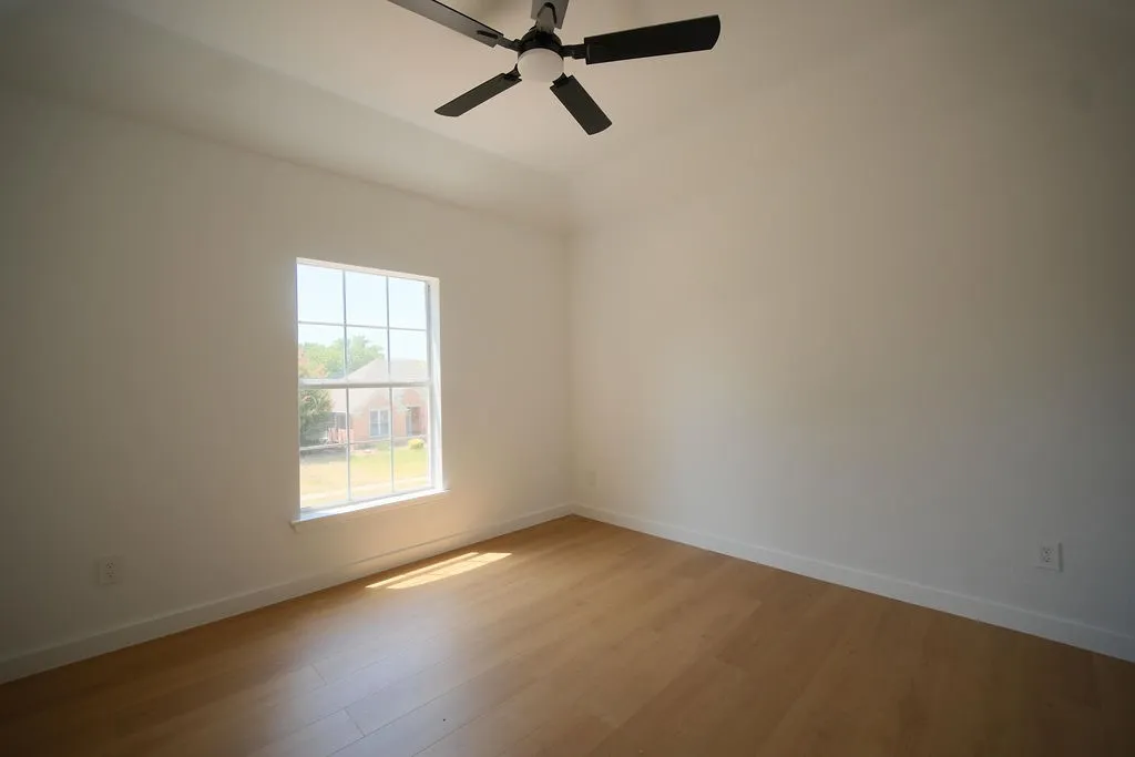 Bright, light-filled bedroom showcasing hardwood flooring, a large double-hung window with grid detailing, and a ceiling fan with integrated lighting