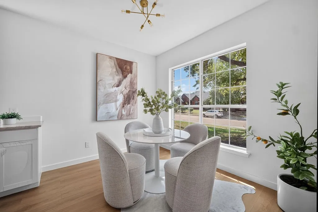 Bright dining area flooded with natural light through expansive multi-pane windows, featuring a round pedestal table, upholstered chairs, and wood-finish flooring