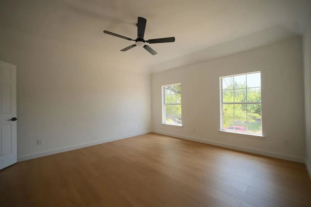 Spacious room flooded with natural light through dual large windows, featuring warm wood-finish flooring and a ceiling fan with brushed metal blades